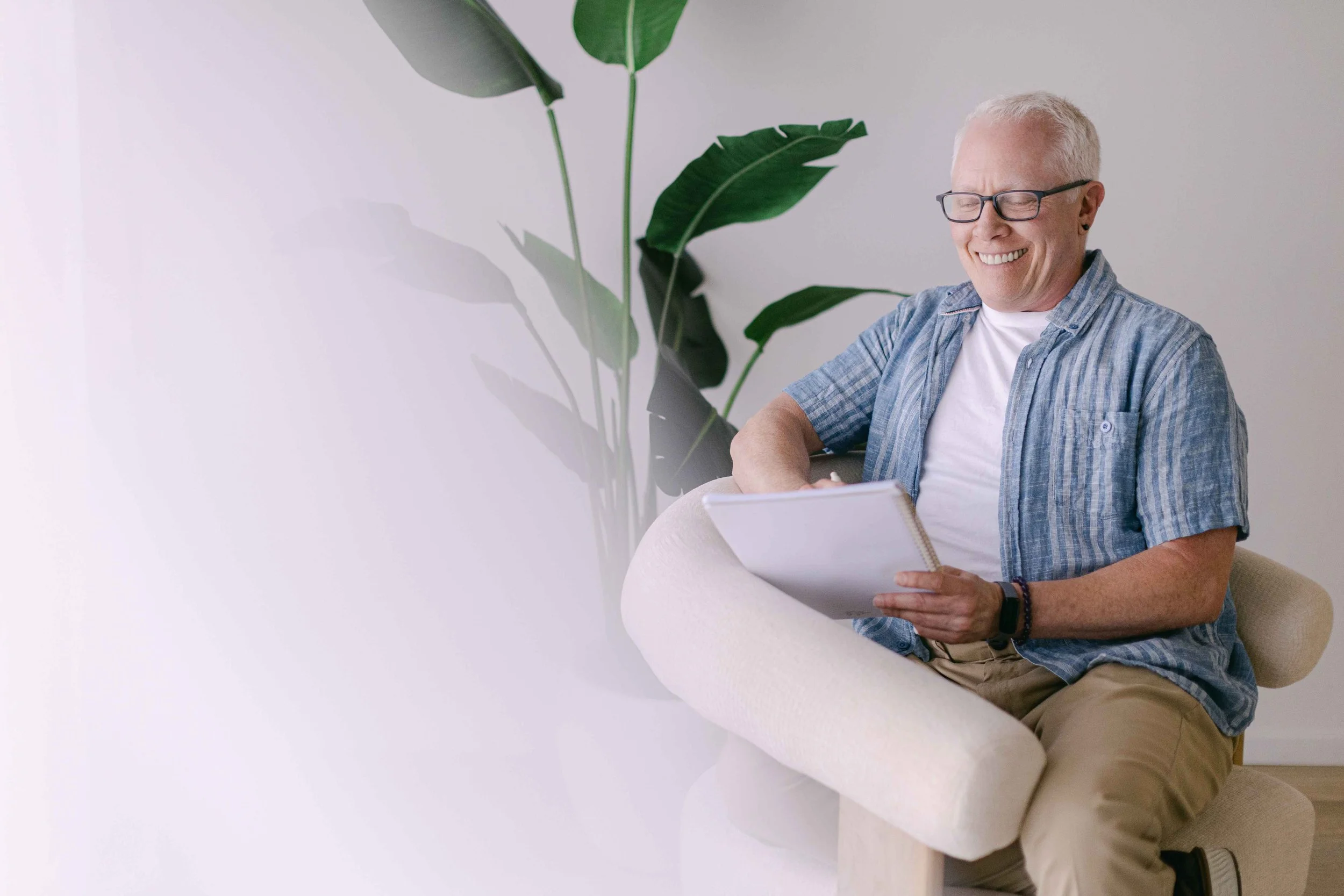 A smiling older man with glasses, wearing a striped blue shirt and khaki pants, sitting on a beige chair in a bright room, holding a notebook, with a large green plant in the background.