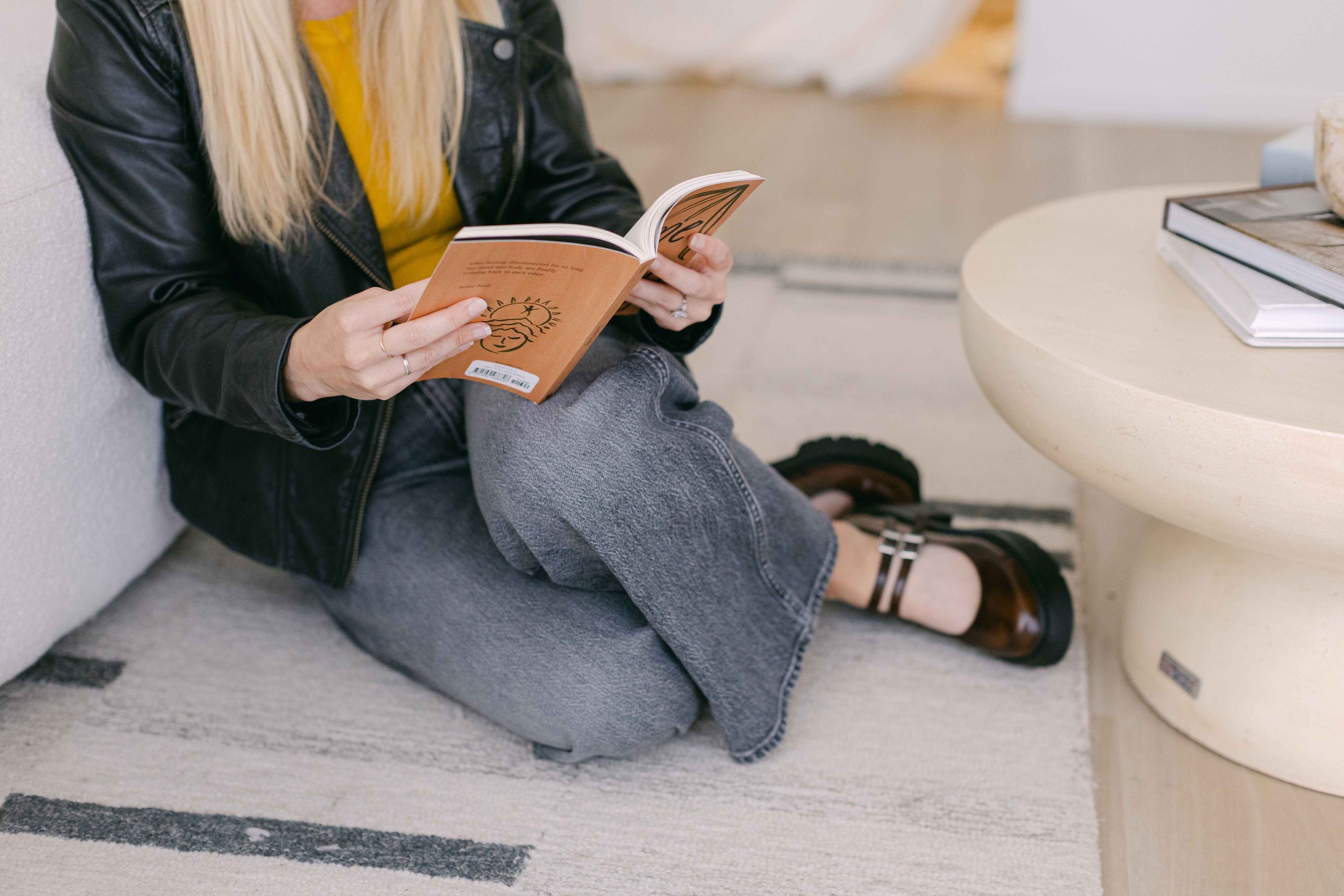 Person sitting on the floor with legs crossed, reading a book, wearing gray jeans, black shoes, a yellow shirt, and a black jacket, near a white table with a stack of books on it.