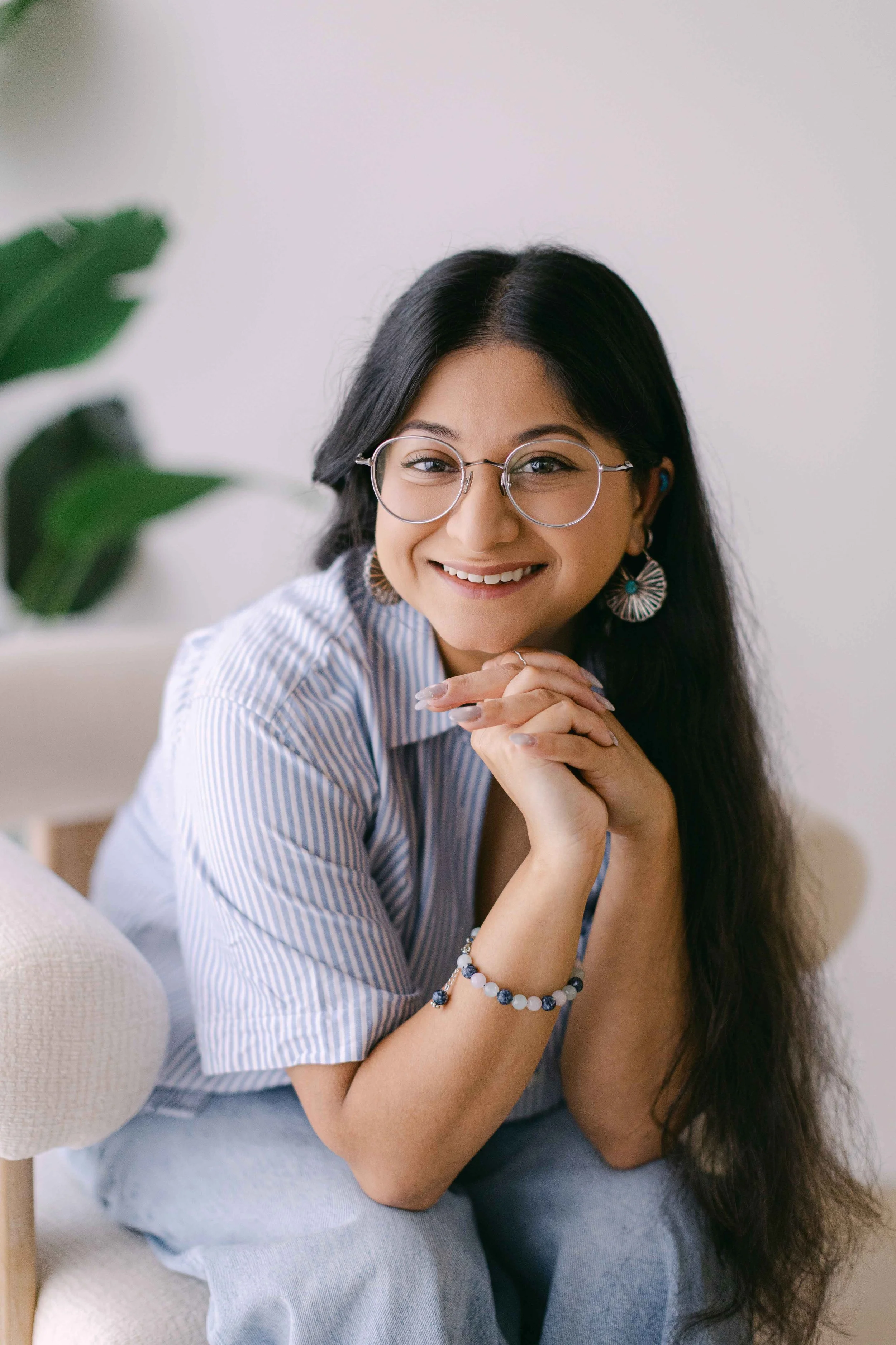A woman with long dark hair, wearing glasses and large earrings, smiling and resting her chin on her clasped hands, sitting indoors with a white wall and a green plant partially visible in the background.