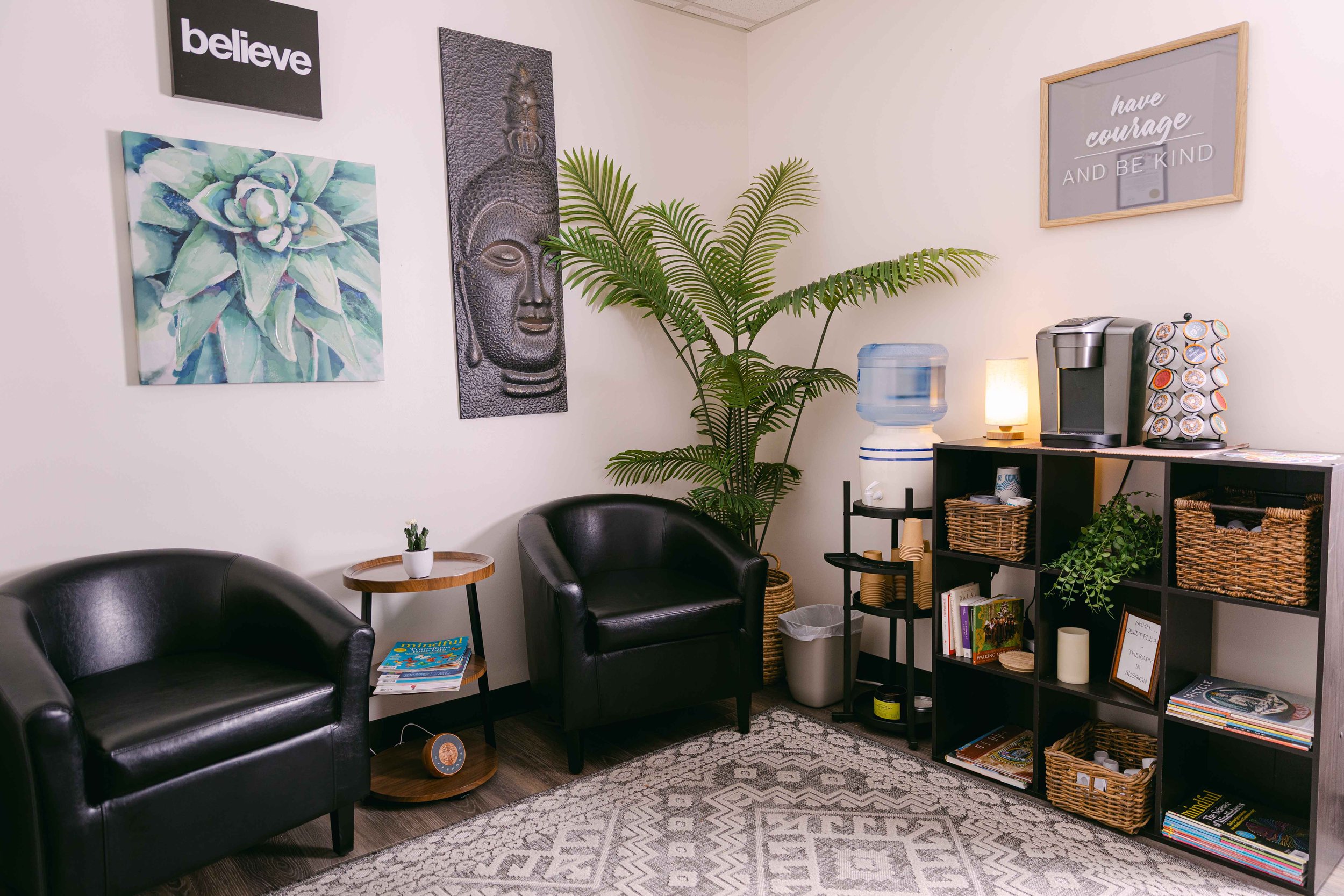 A cozy waiting or lounge area with two black armchairs, a small wooden side table with a potted plant, and a patterned rug. The wall has several decorative pieces including a black sign with