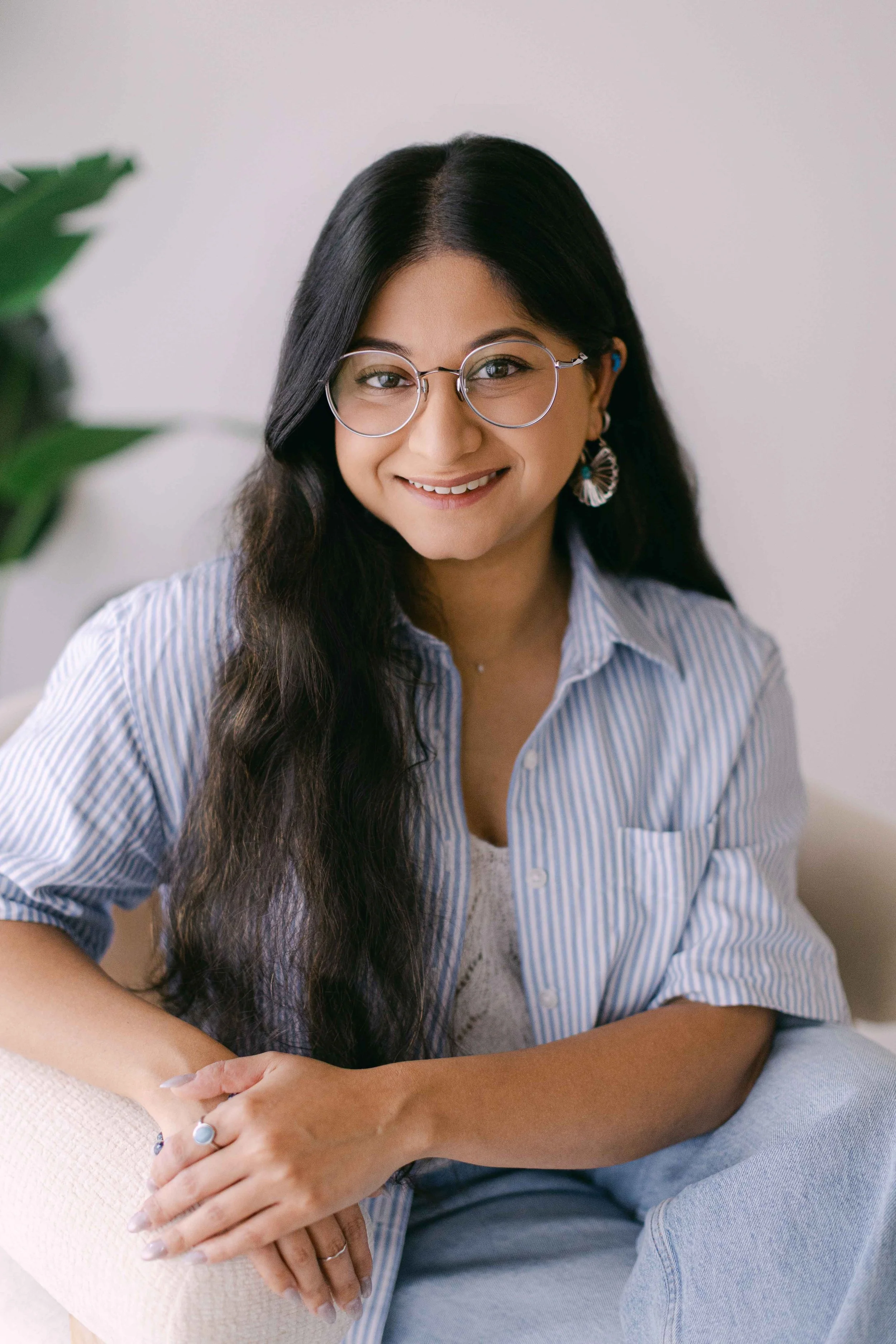 A woman with long dark hair, glasses, and earrings, smiling while sitting on a beige chair with a white wall background.