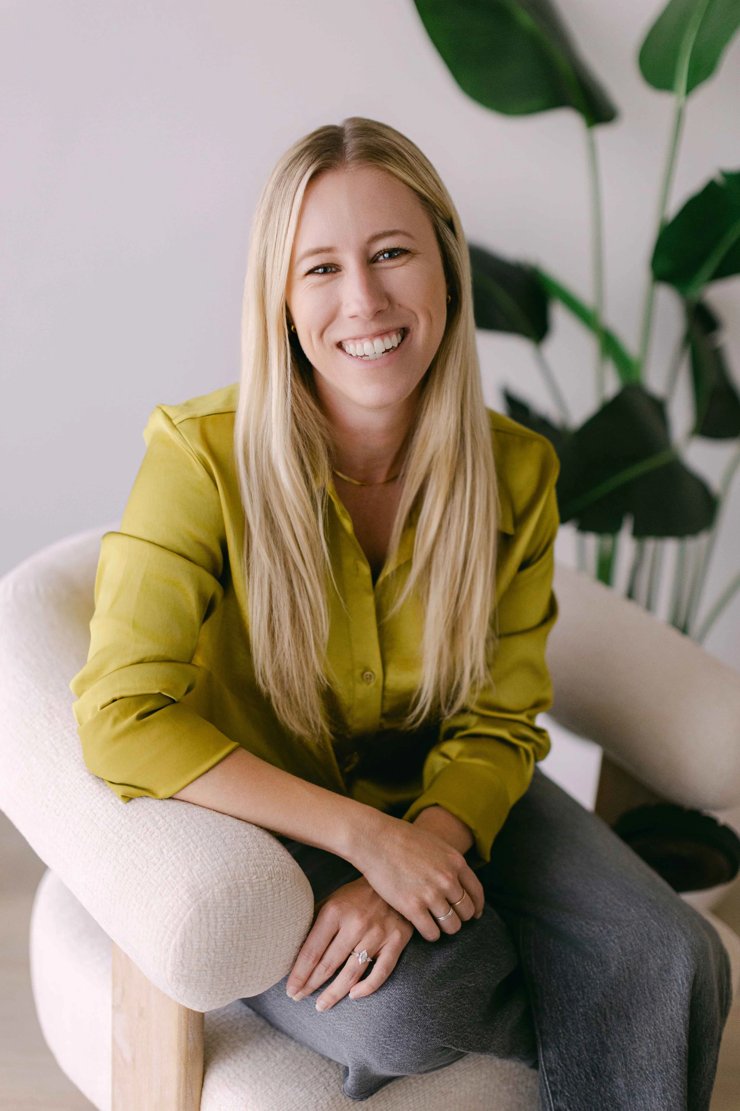 A smiling woman with long blonde hair, wearing a yellow blouse, sitting on a beige armchair with her hands resting on her lap, in front of a white wall with a tall green plant in the background.