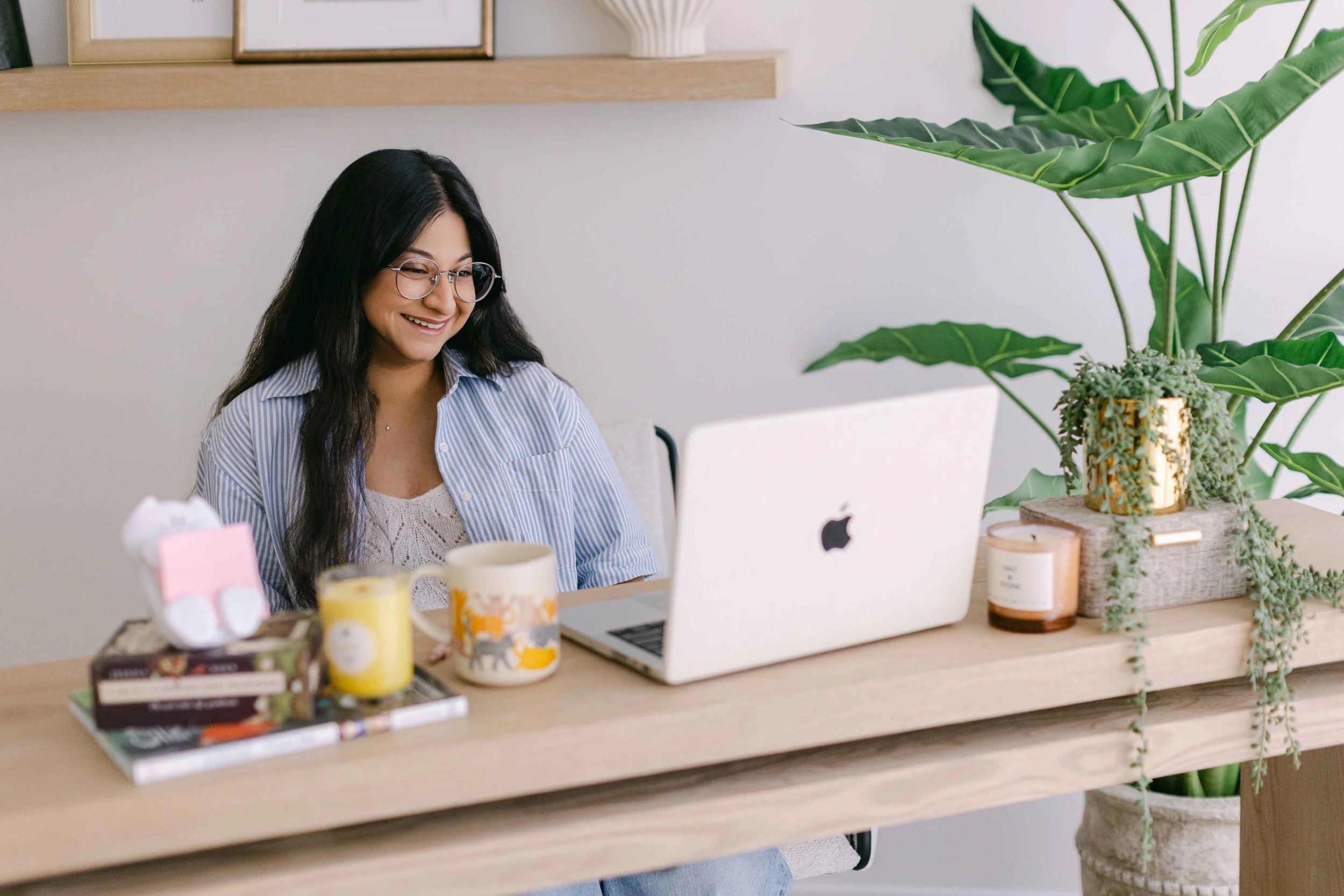 A woman with long black hair and glasses smiling while working at her desk with a computer, surrounded by potted plants and office supplies.