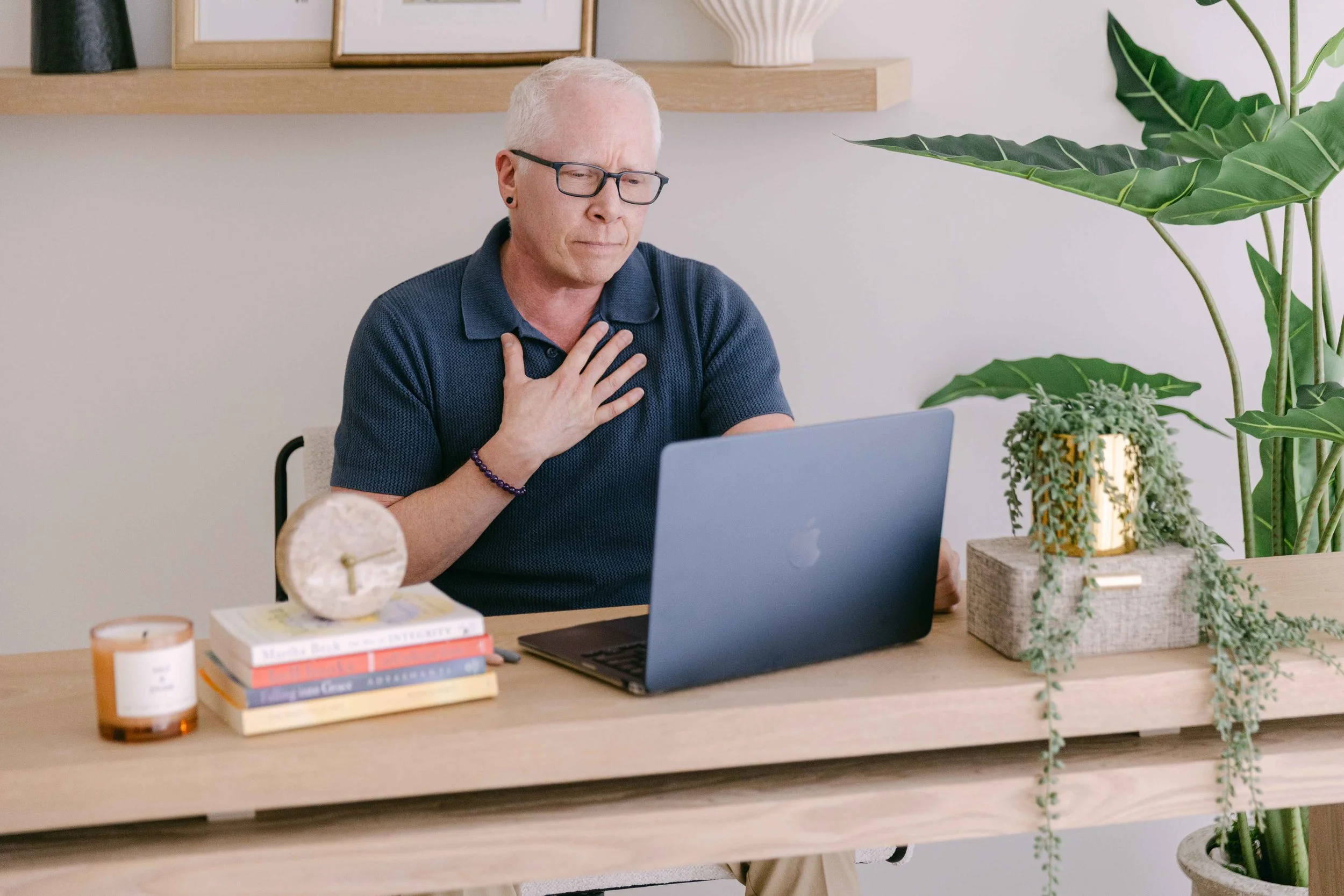 A man with white hair, glasses, and earrings is sitting at a desk with a laptop, holding his chest and appearing to be in discomfort.