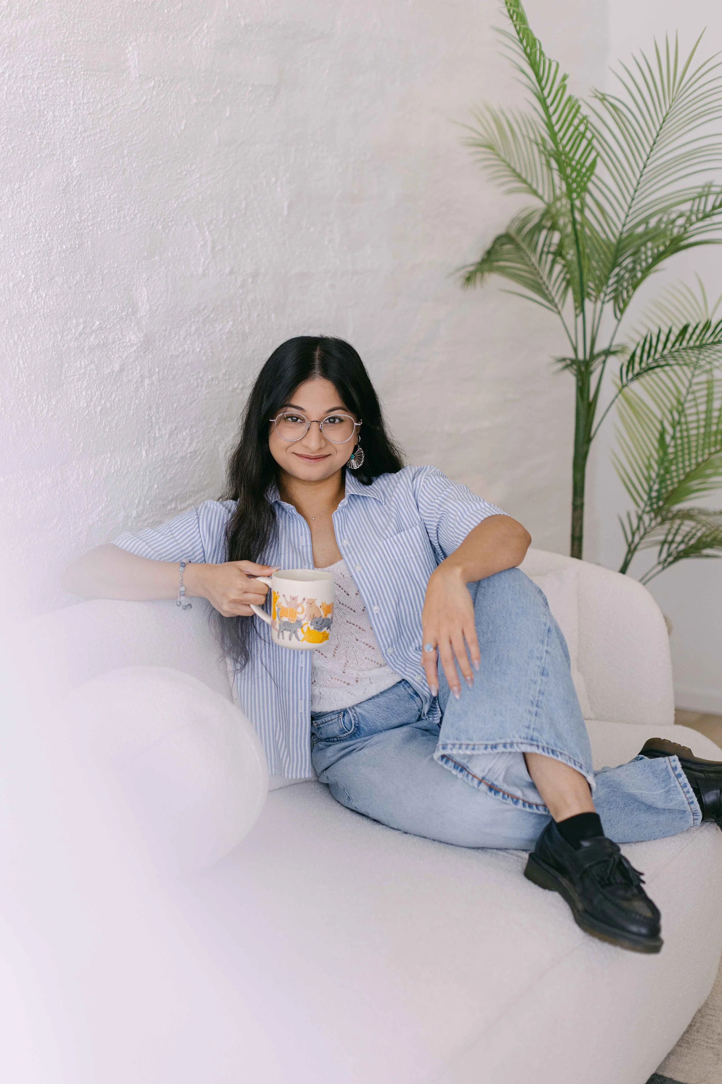 Young woman with glasses sitting on a white sofa, holding a colorful mug, wearing a striped shirt, jeans, and black shoes, with a large green plant in the background.