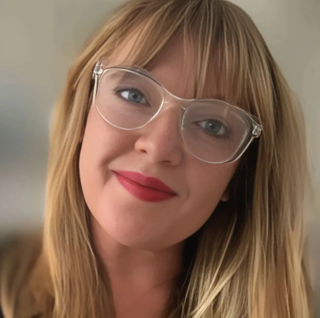 Close-up of a woman with long blonde hair, wearing clear glasses and red lipstick, smiling softly.