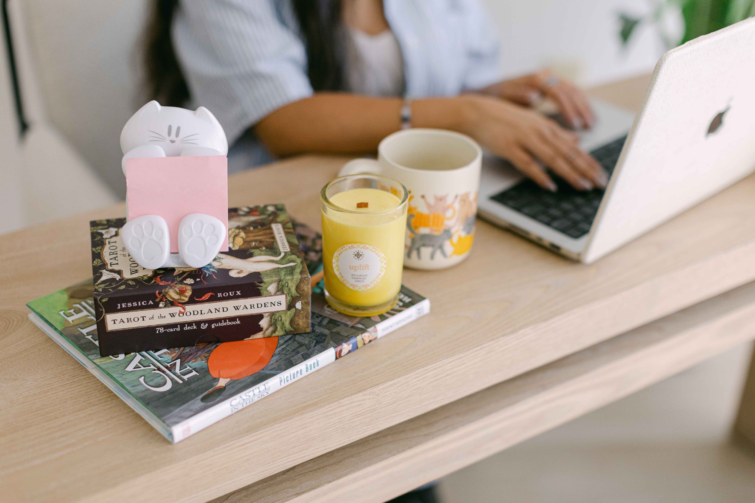 A desk with a tarot card deck, books, a yellow candle, a mug with elephant prints, a white cat-shaped object with a pink note, and a person typing on a silver laptop.