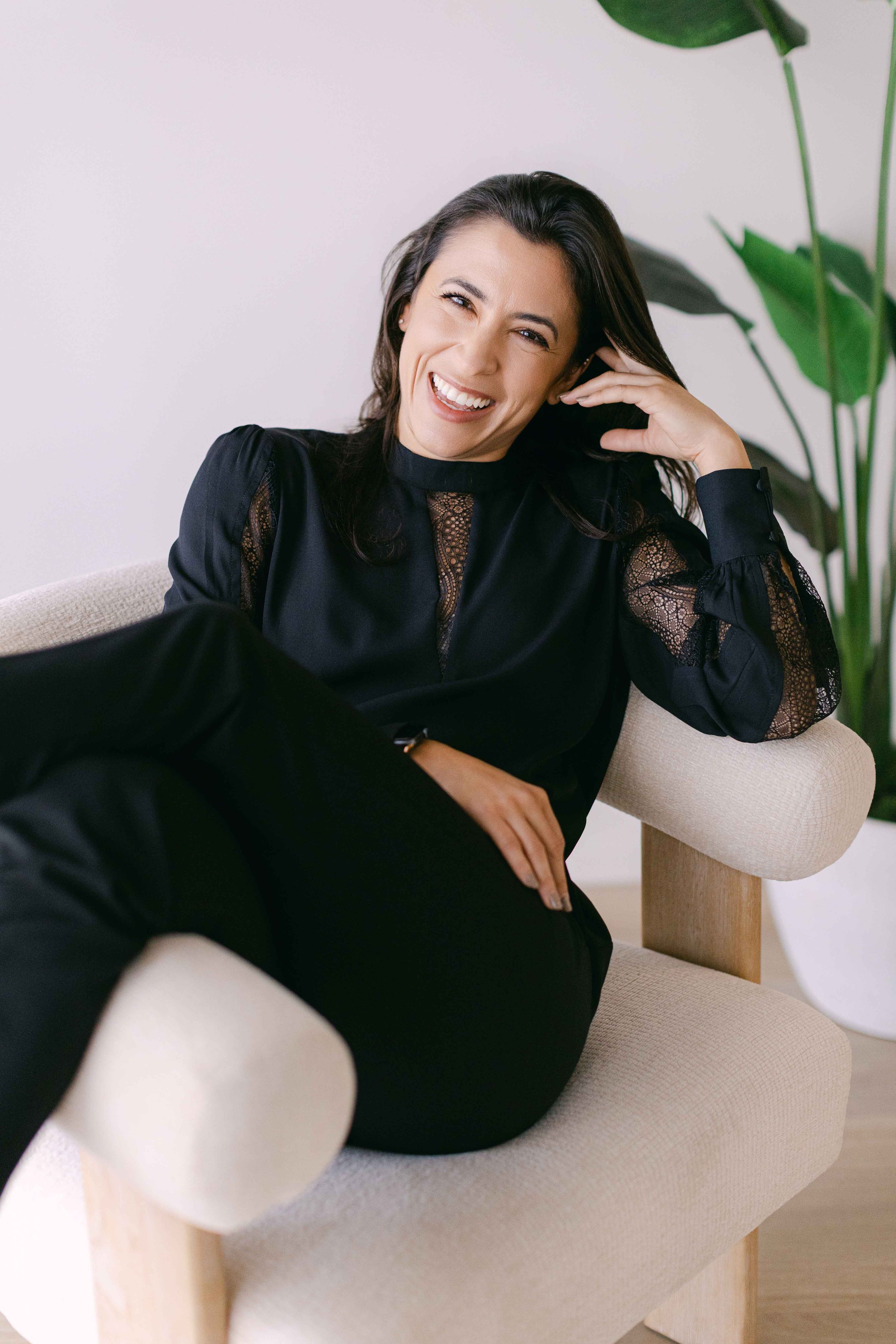 A woman with dark hair, smiling and laughing, sitting on a light-colored armchair with her hand resting on her head. She is wearing a black, long-sleeved blouse with lace accents on the sleeves and neckline. There is a large green plant with broad leaves in the background.