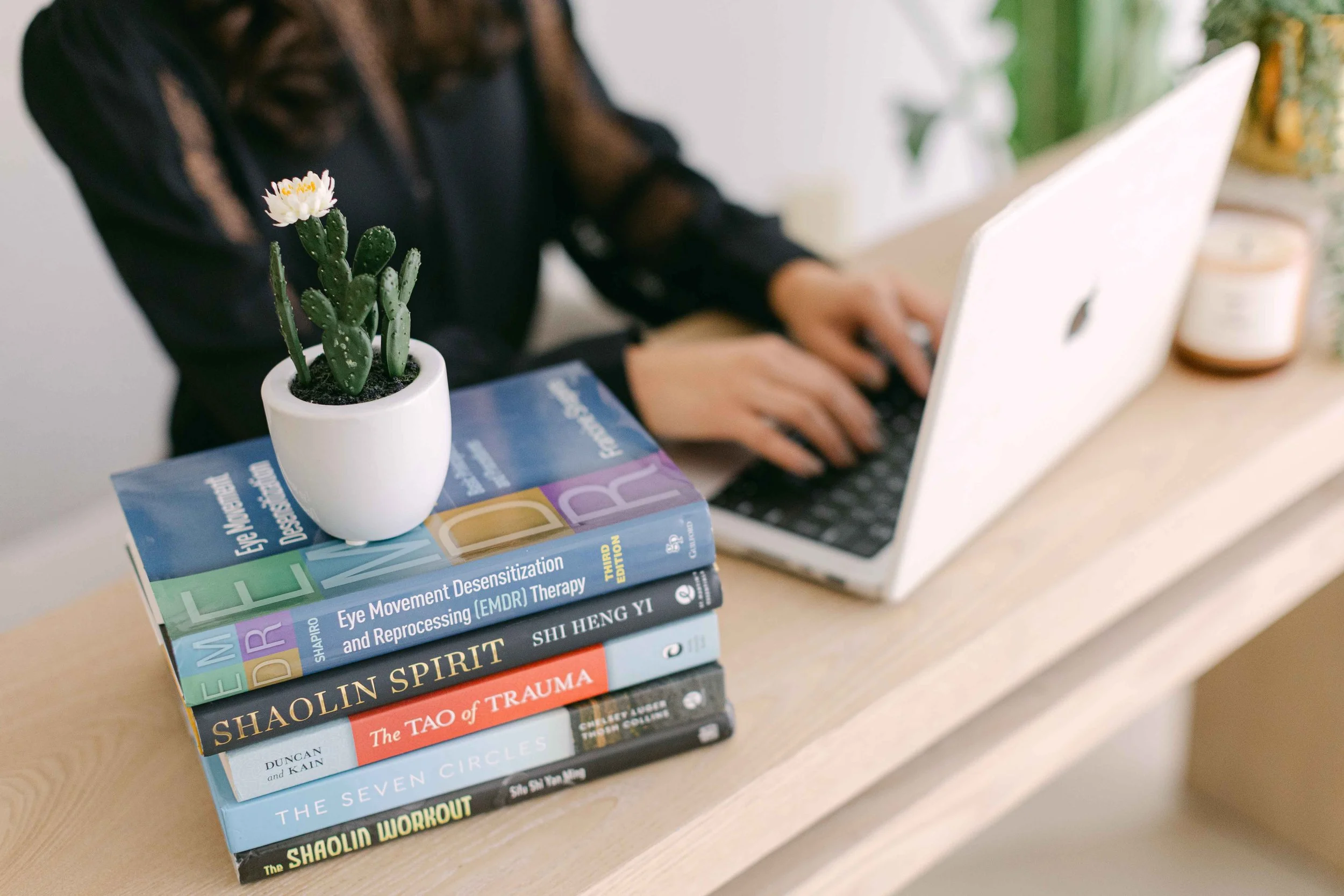 Stack of books on a desk with a small potted cactus plant in a white pot on top of the books. The books cover topics like eye movement processing, trauma, and spirituality. In the background, a person is using a laptop.
