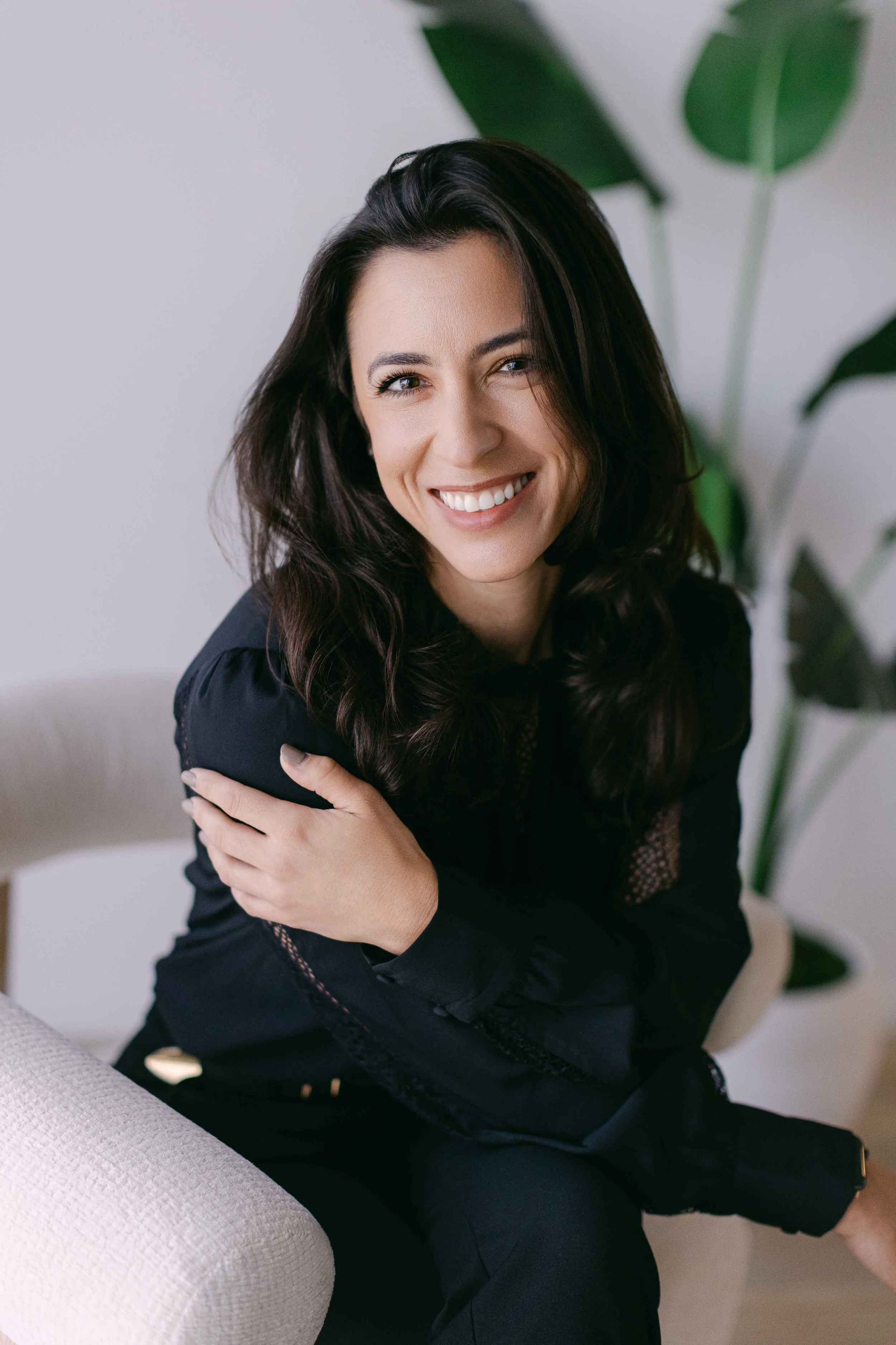 Smiling woman with dark hair, sitting on a beige couch, wearing a black top, with a large green leafy plant in the background.
