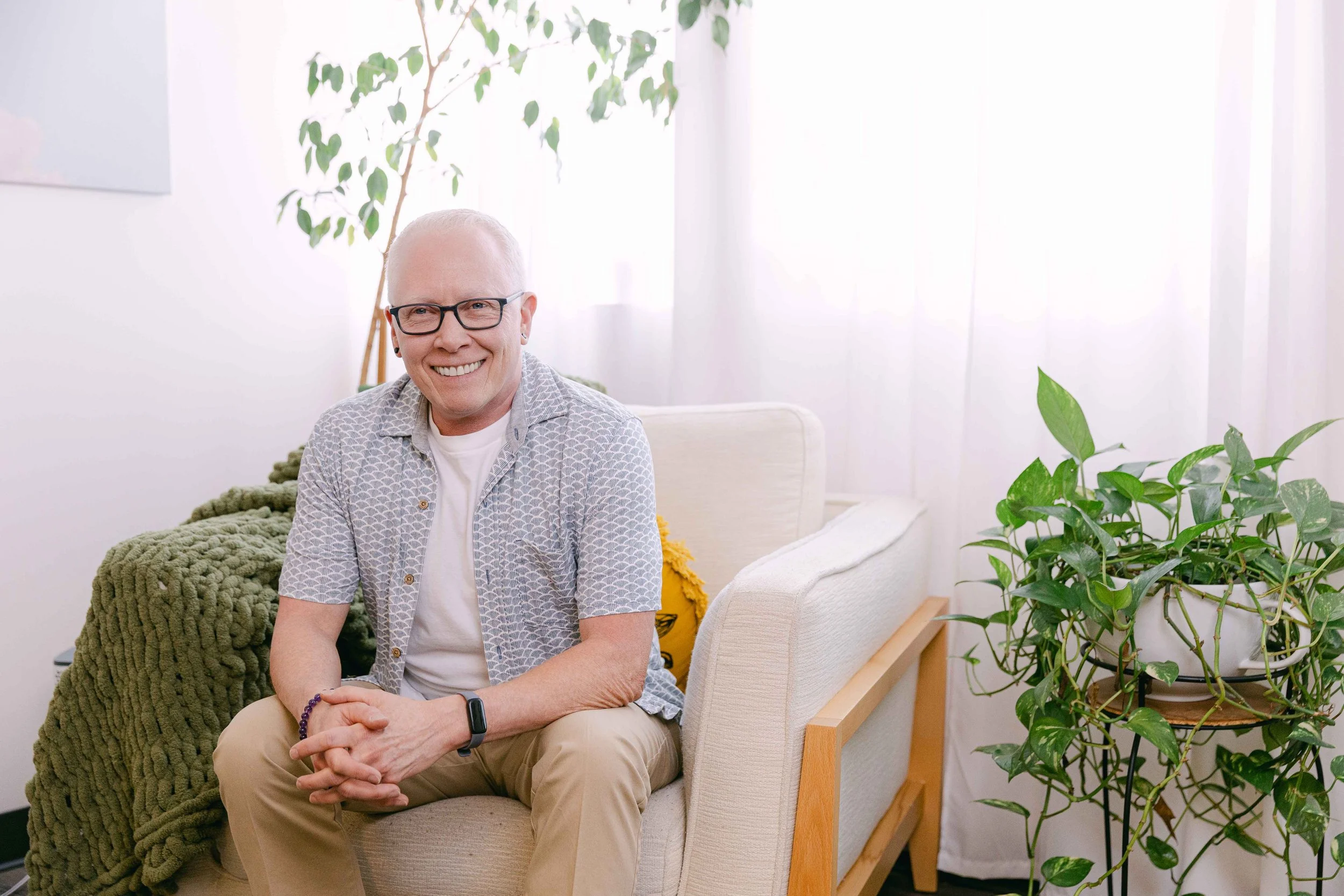 A smiling person with glasses and short white hair sitting on a beige chair in a well-lit room with houseplants and white curtains.