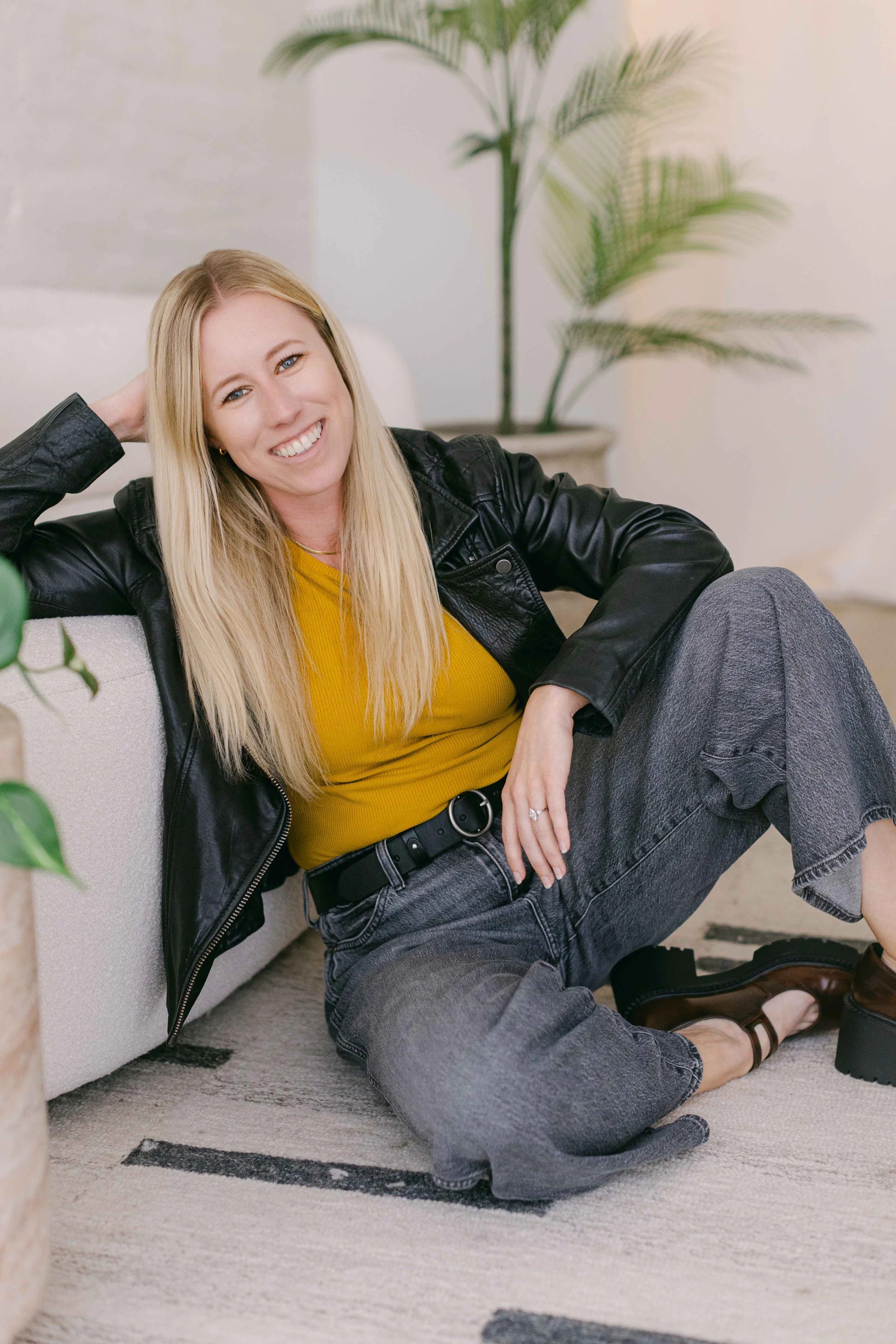 A woman with long blonde hair smiling and sitting on the floor with her back against a beige couch. She is wearing a black leather jacket, a yellow top, and gray jeans. The woman has one hand resting behind her head and the other on her knee. There is a large potted plant with long, feather-like leaves in the background. The setting appears to be a cozy, well-lit living room.