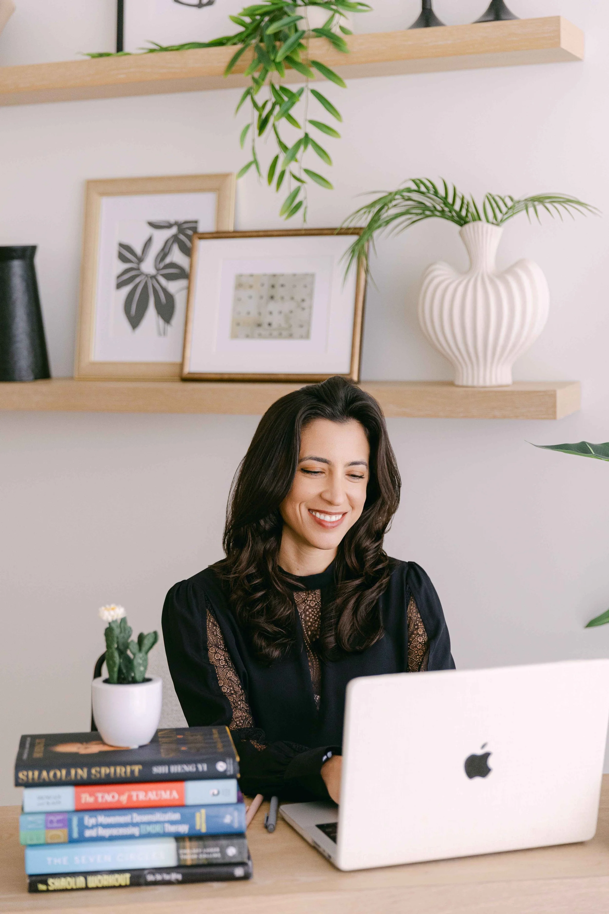 A woman with long dark hair smiling at her workspace, sitting in front of a white Apple laptop. On her desk, there is a stack of books, a notepad, and a small cactus plant in a white pot. The background shows a white wall with floating shelves holding framed art, plants, and decorative items.