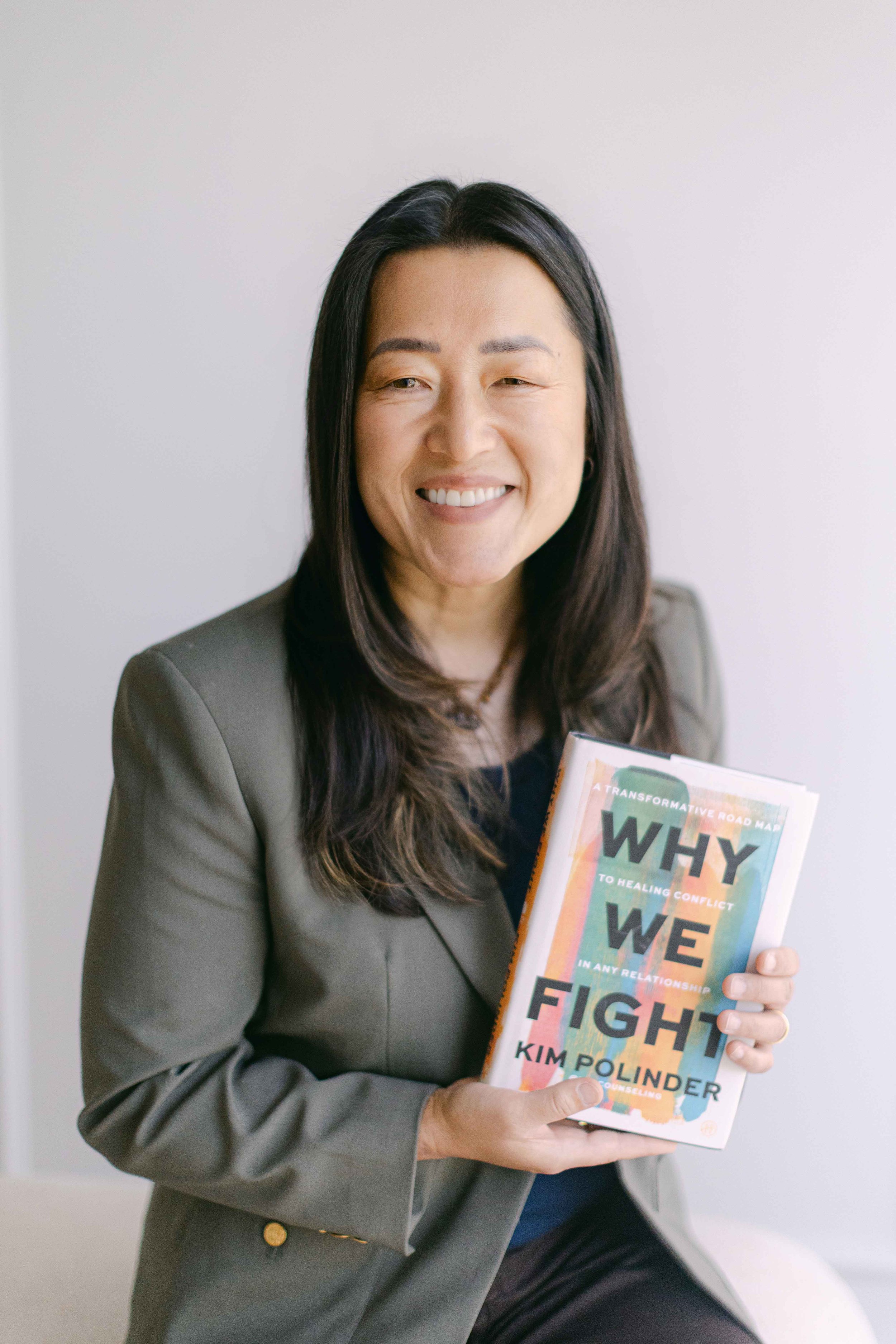 A woman with long dark hair, wearing a gray blazer and black top, smiling and holding a colorful book titled 'Why We Fight' by Kim Polinder, against a plain white background.