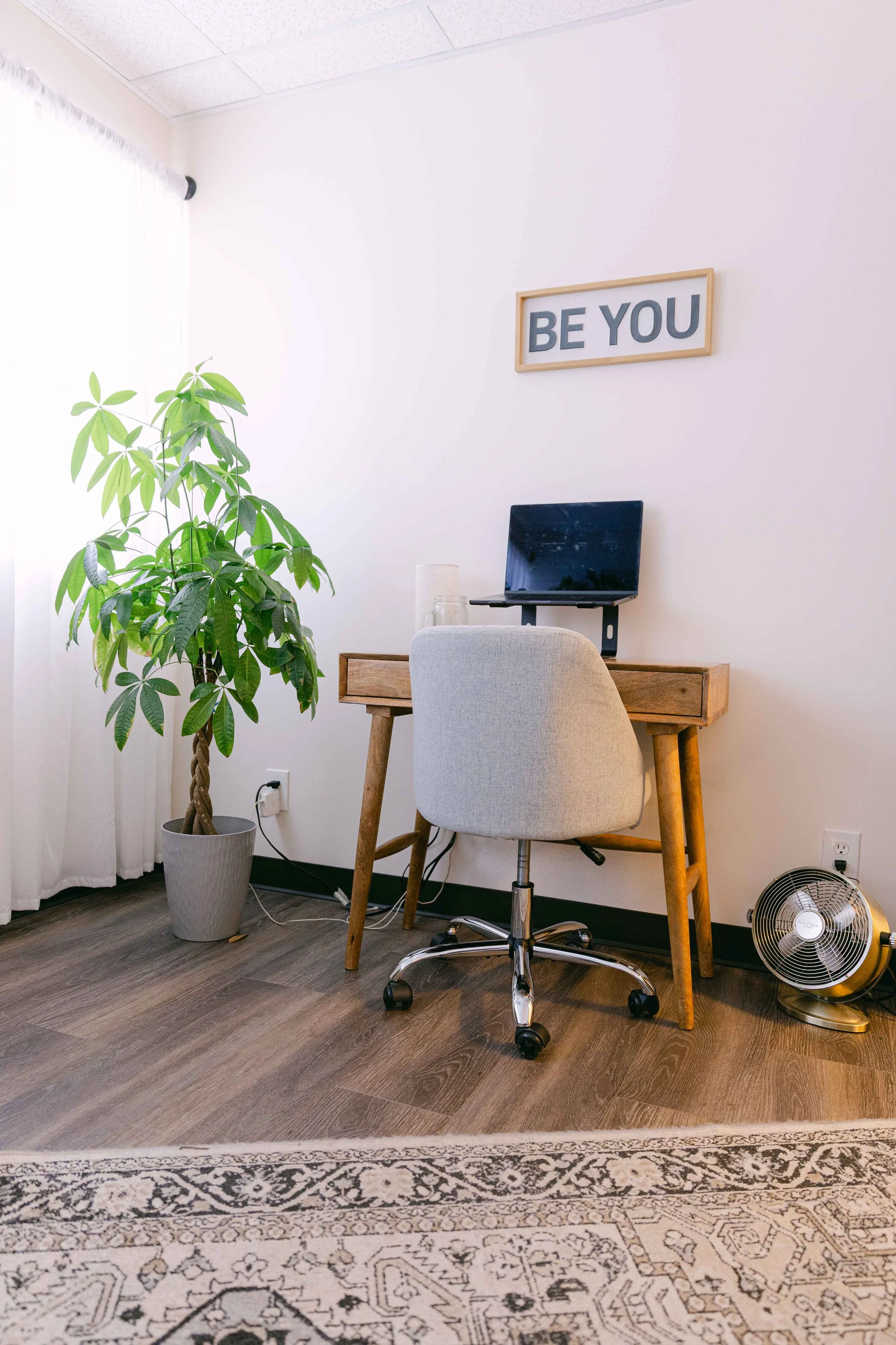 A small workspace with a wooden desk and a beige office chair. On the desk, there is a computer monitor, and behind it, a framed sign with the words "BE YOU". To the left, there's a tall potted plant with large green leaves. To the right, on the floor, there's a small fan. The room has wood flooring, white walls, a patterned area rug, and sheer curtains on the window letting in natural light.