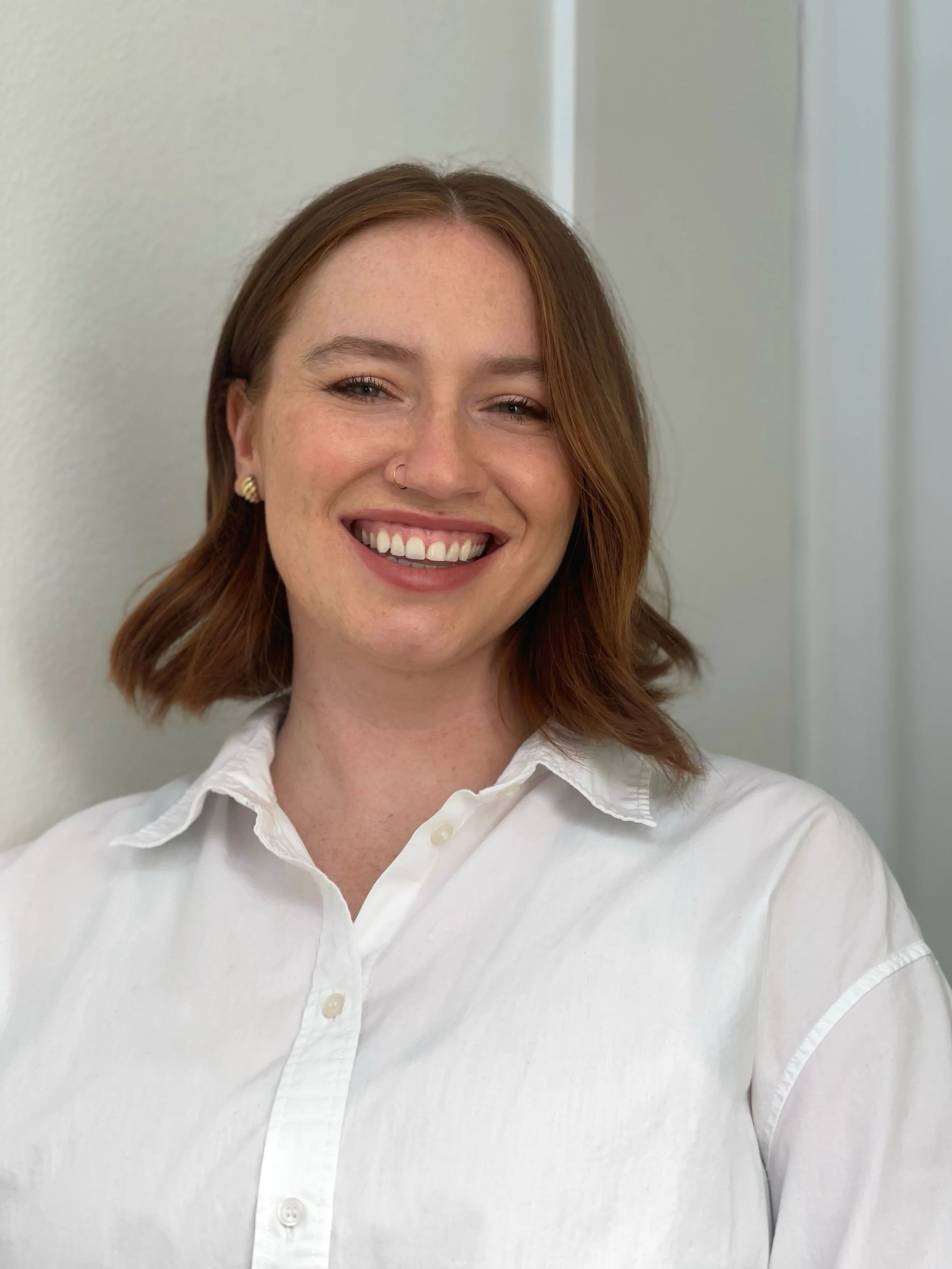 Close-up of a smiling woman with red hair, wearing a white shirt and small earrings, standing against a plain wall.