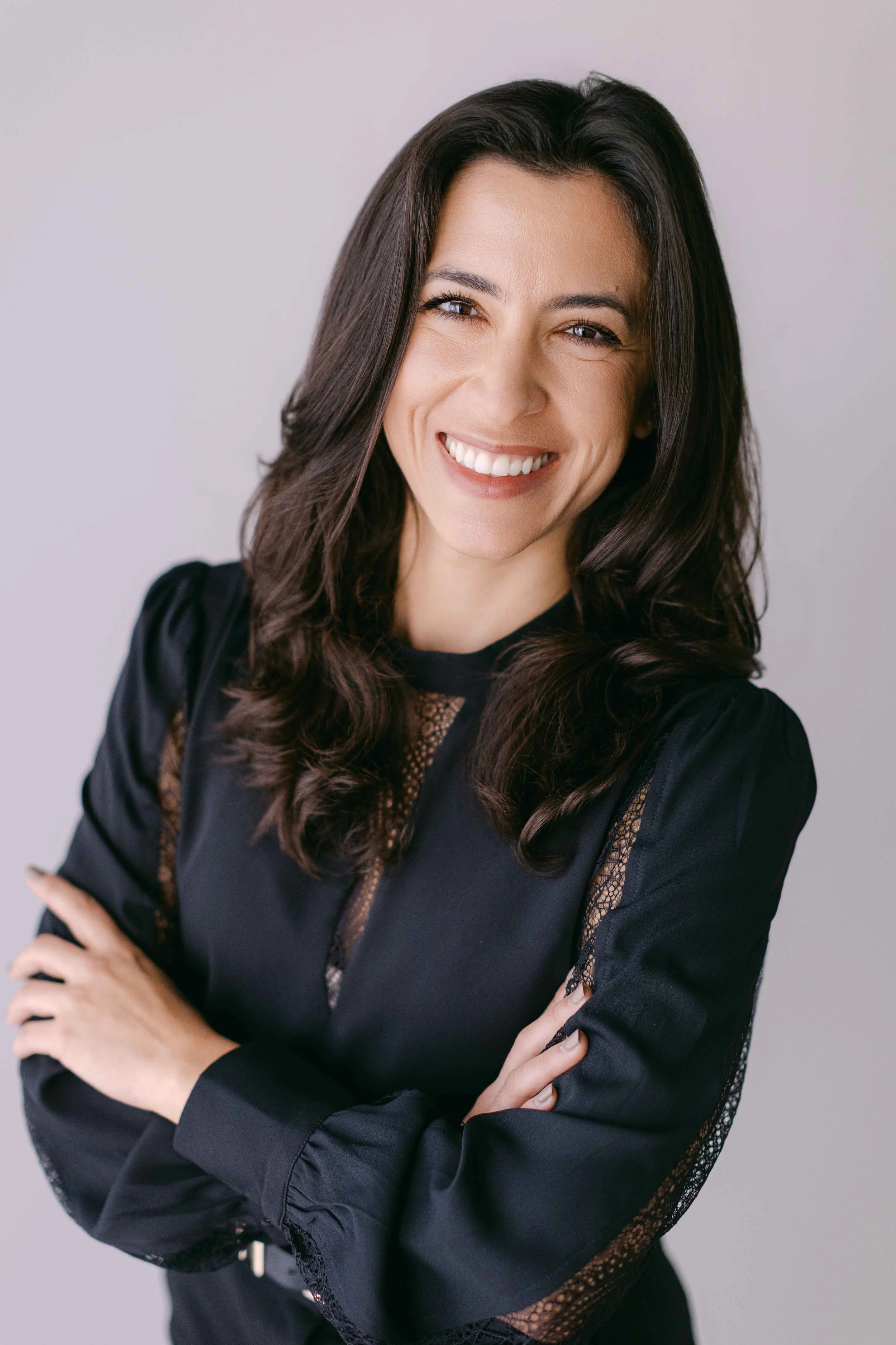 A woman with long dark hair, smiling, wearing a black top with lace details, crossing her arms in front of a plain light background.