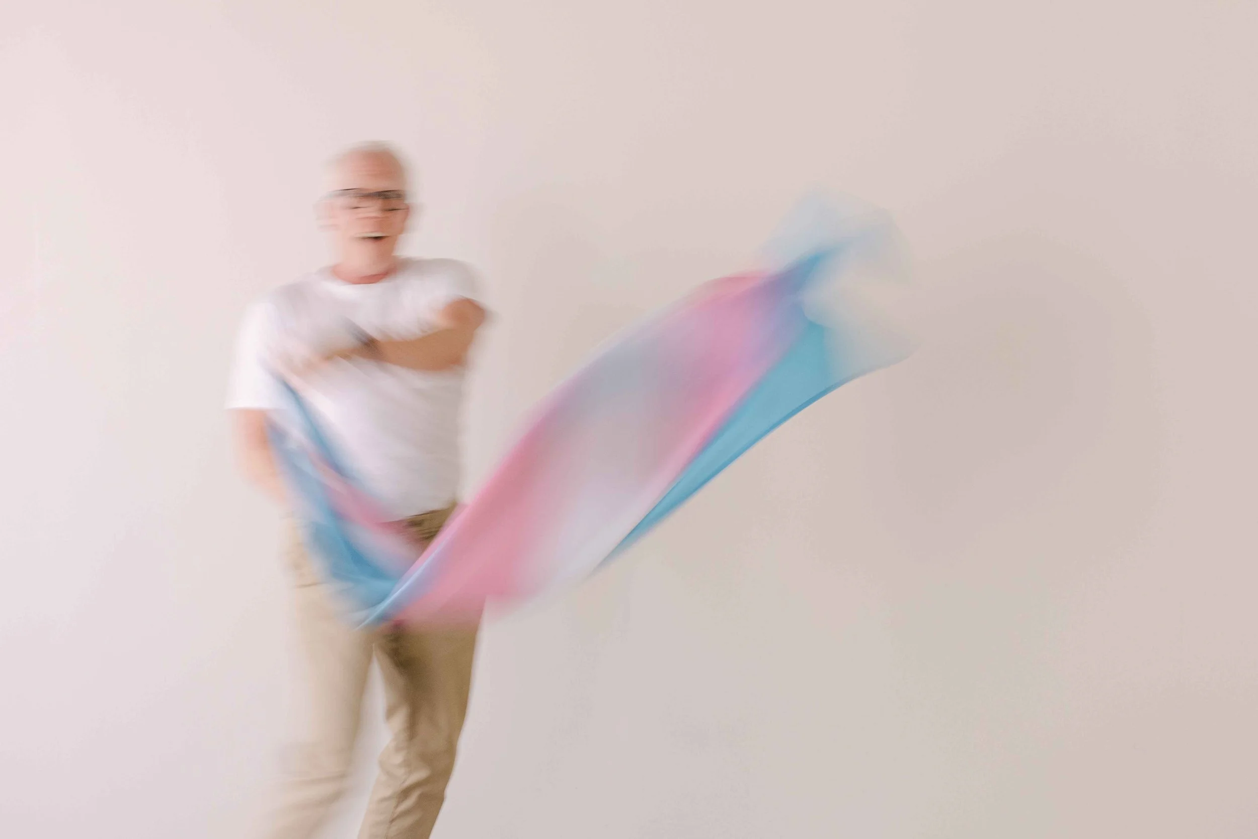 A man with glasses and a white shirt is playing with a colorful fabric in front of a plain light-colored background.
