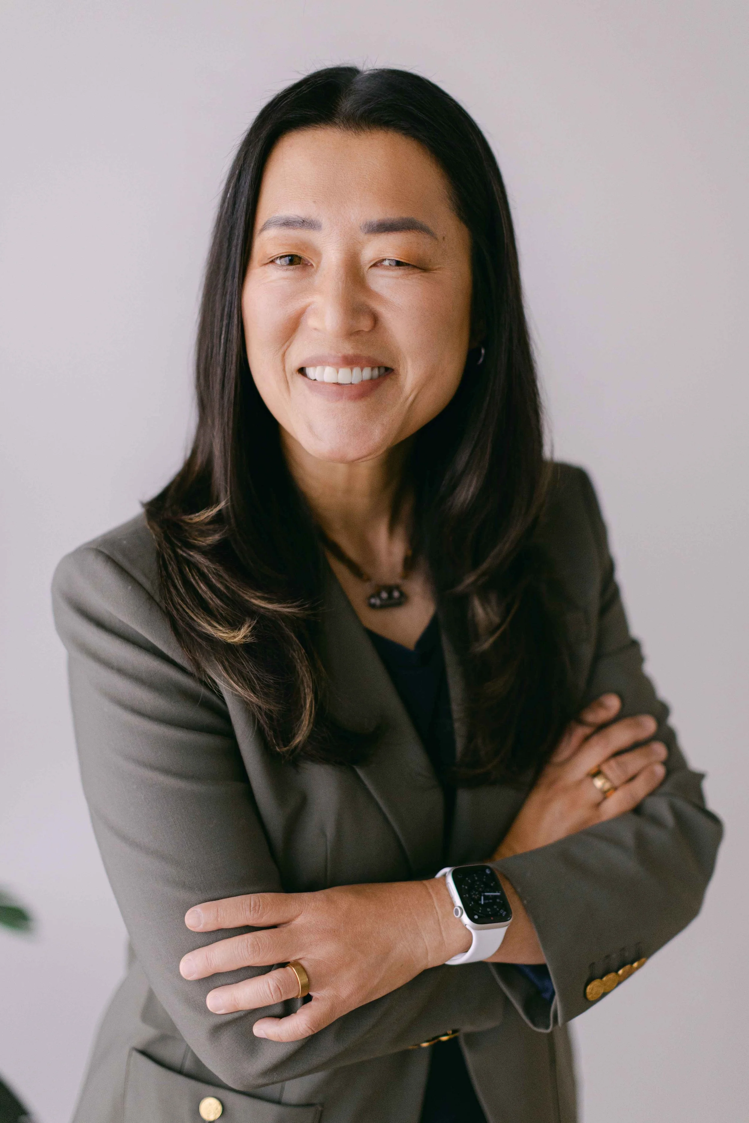 A woman with dark hair and a close shave, smiling with anime eyes, wearing a gray blazer and a smartwatch, standing against a plain background with arms crossed.