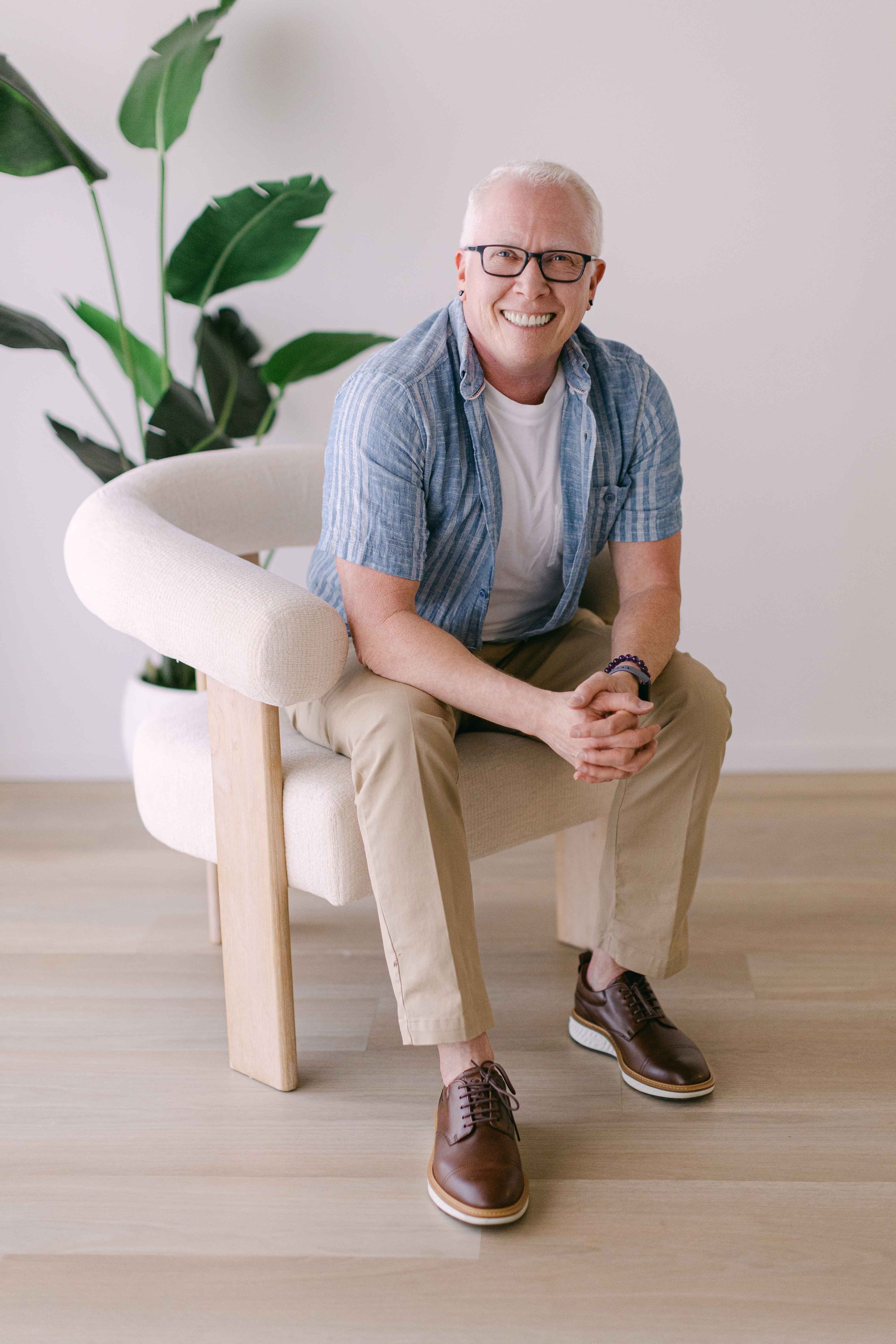 A man with glasses and short white hair smiling, sitting on a light-colored armchair with wooden legs, wearing a blue plaid shirt, beige pants, and brown shoes, with a large leafy plant in the background against a plain white wall.