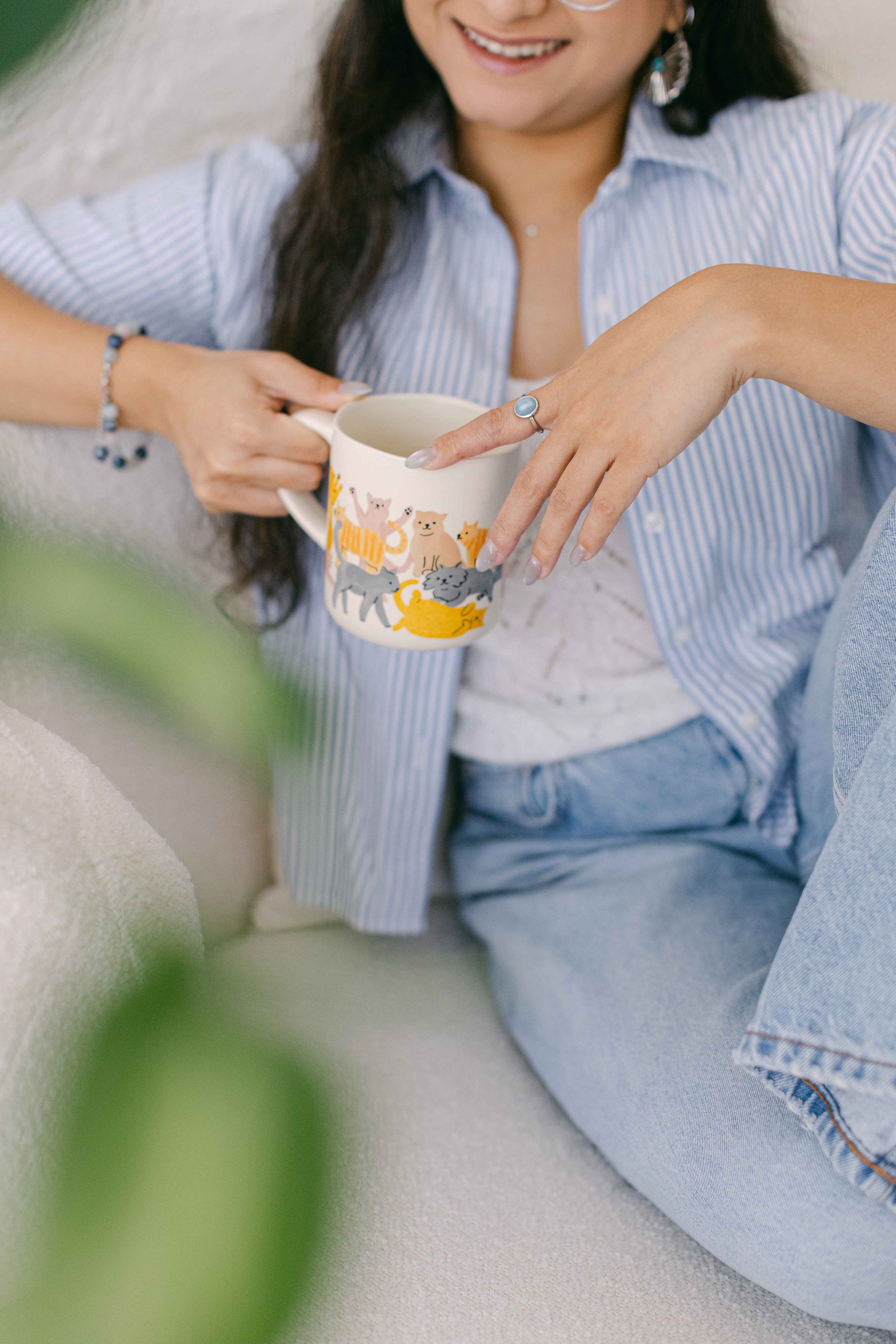A woman with long dark hair smiling and wearing a blue and white striped shirt, accessories including earrings, rings, and bracelets, holding a white mug decorated with colorful animal illustrations, sitting on a light-colored couch.