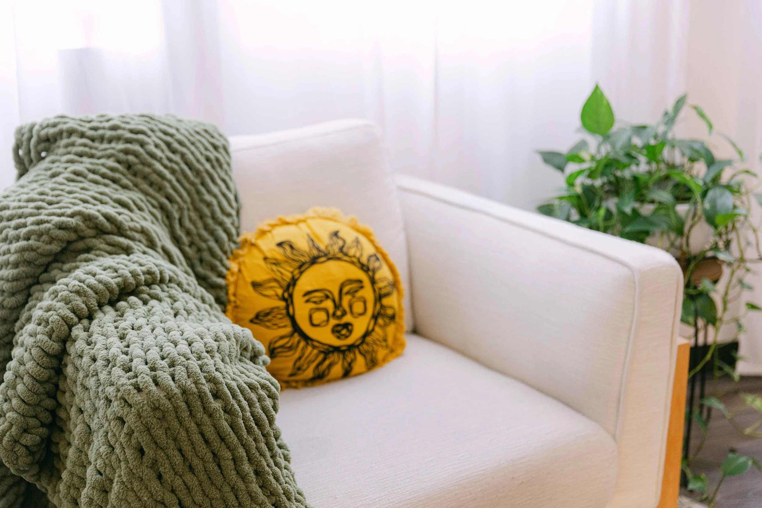 A cozy white sofa decorated with a chunky green blanket and a yellow pillow with a sun and woman's face illustration, next to a green leafy houseplant.