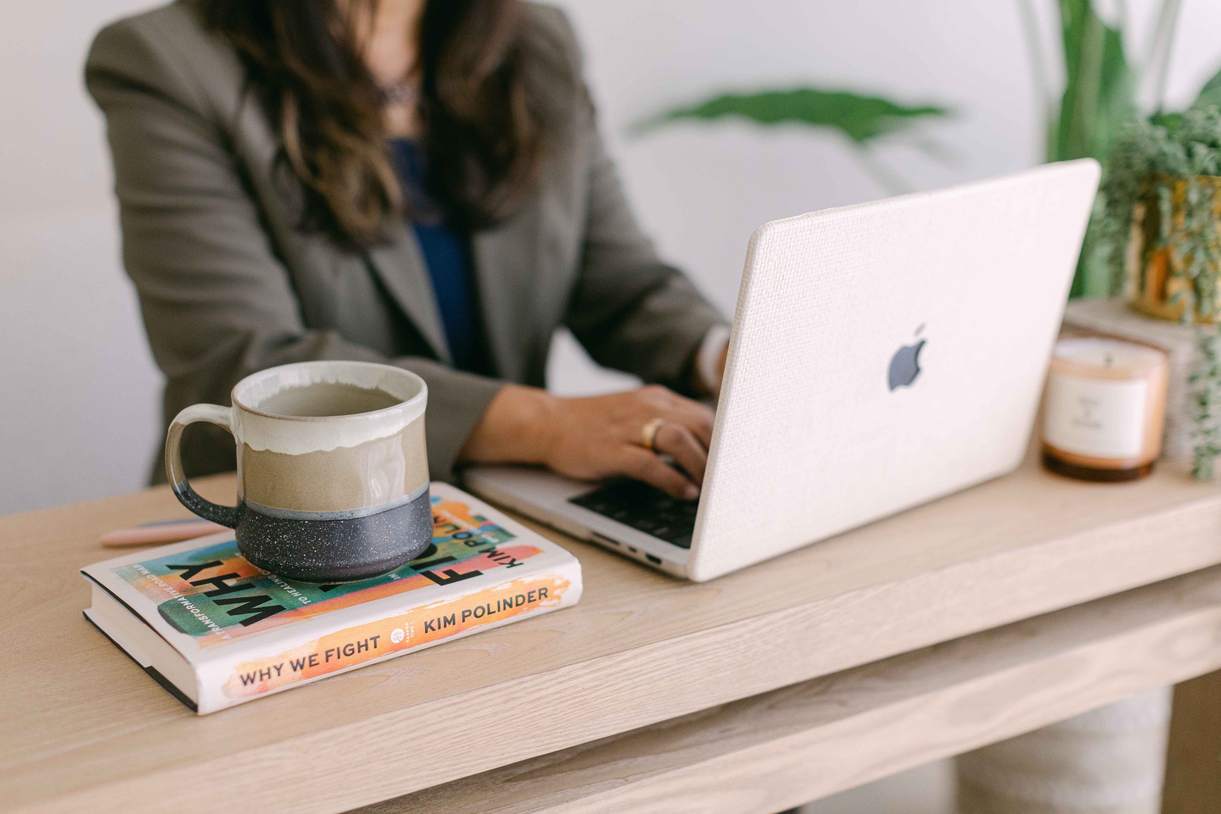 A person working on a laptop at a desk with a cup of coffee and two books, one titled 'Why We Fight' by Kim Polinder, on top.