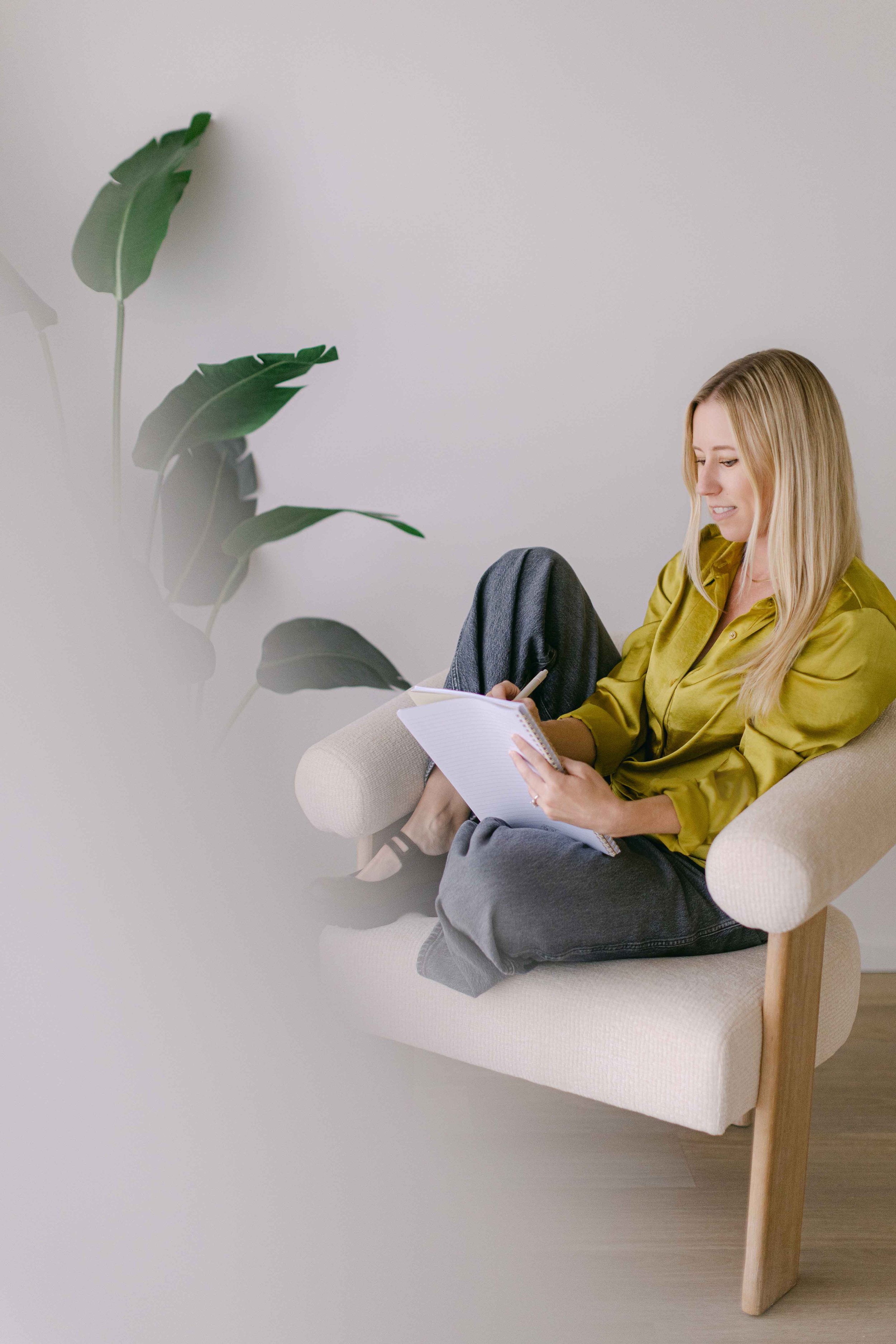 A woman with blonde hair sitting on a cream-colored armchair, smiling and writing in a notebook, wearing a yellow blouse and gray pants, next to a potted plant with large green leaves against a plain white wall.