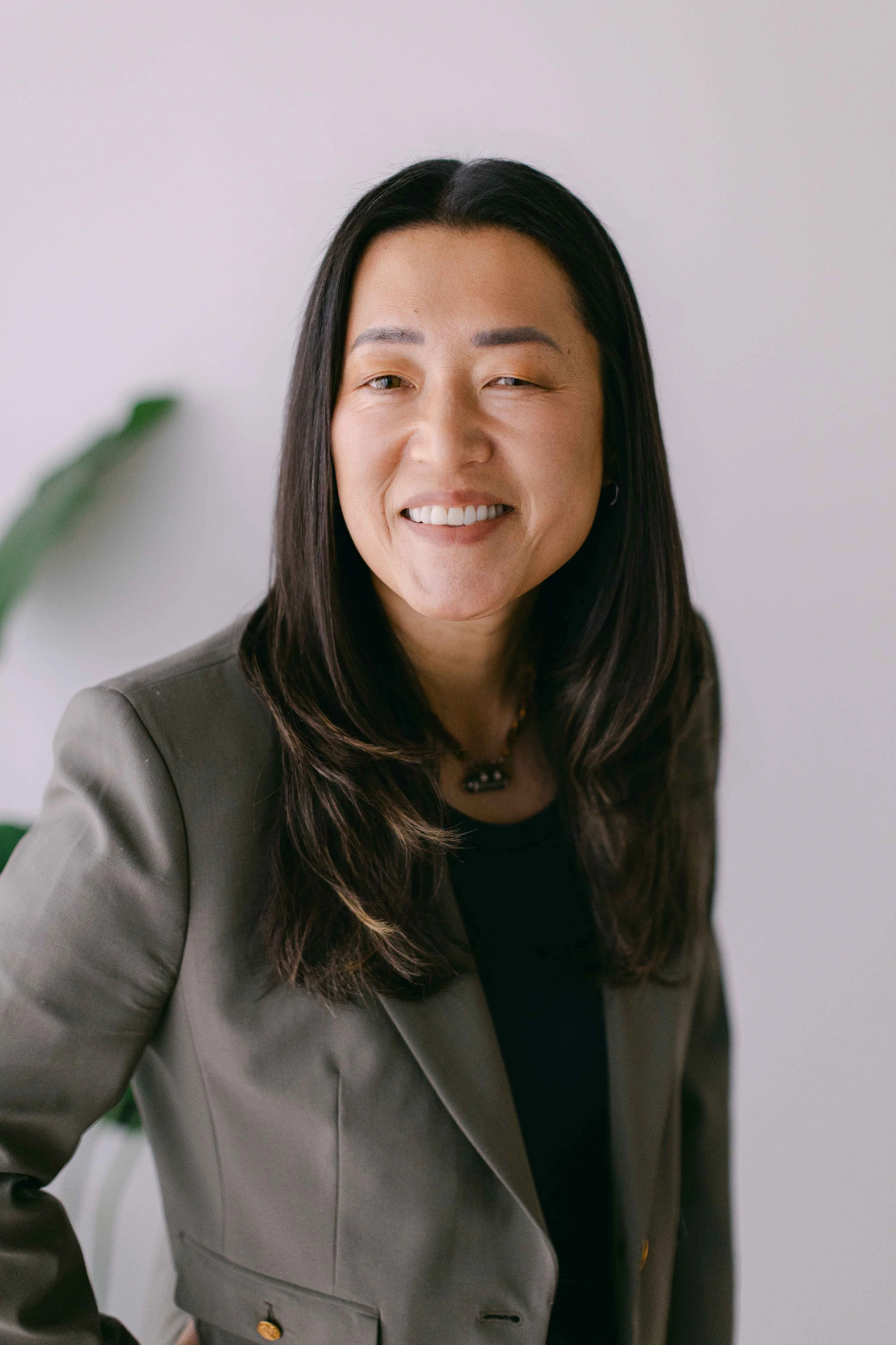 A woman with long, dark hair wearing a gray blazer and black shirt, smiling, with a white wall background and a blurred green plant in the foreground.