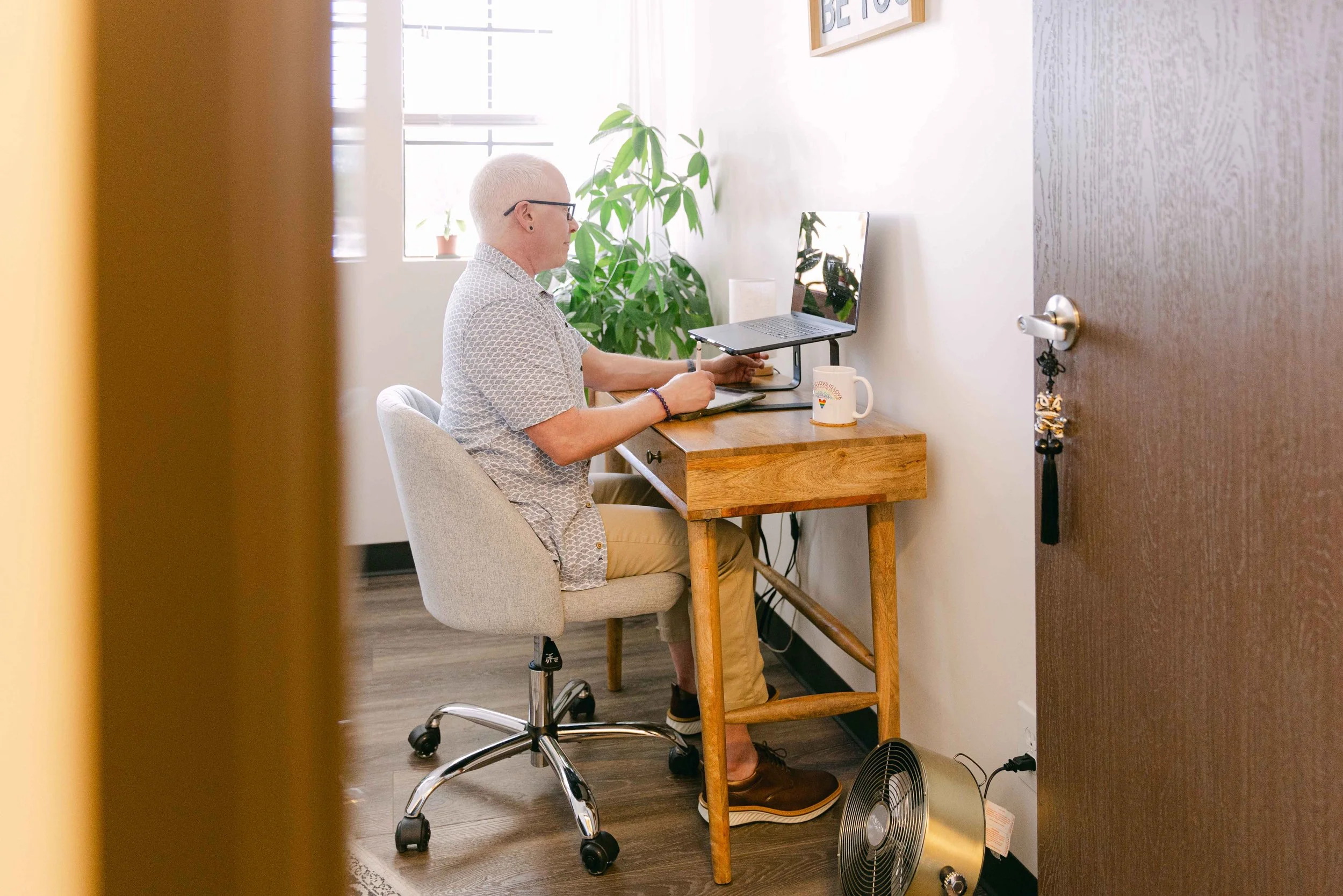 A person with short white hair and glasses working at a wooden desk in a bright room, with a laptop on a stand, a mug, and a large green plant in the background.