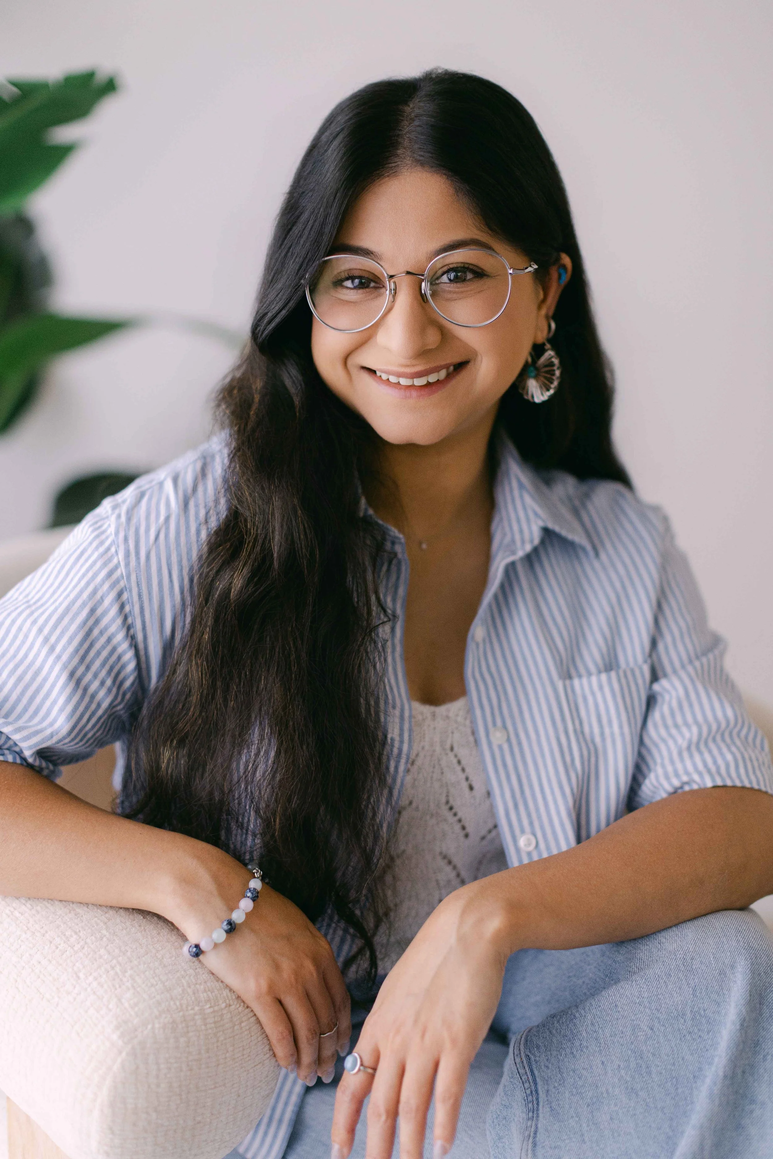 A young woman with long dark hair and glasses smiling while sitting on a beige sofa with a woman with light skin, gray top, and striped blue and white shirt. She is wearing jewelry, including earrings and bracelets, and a white dress underneath her shirt.