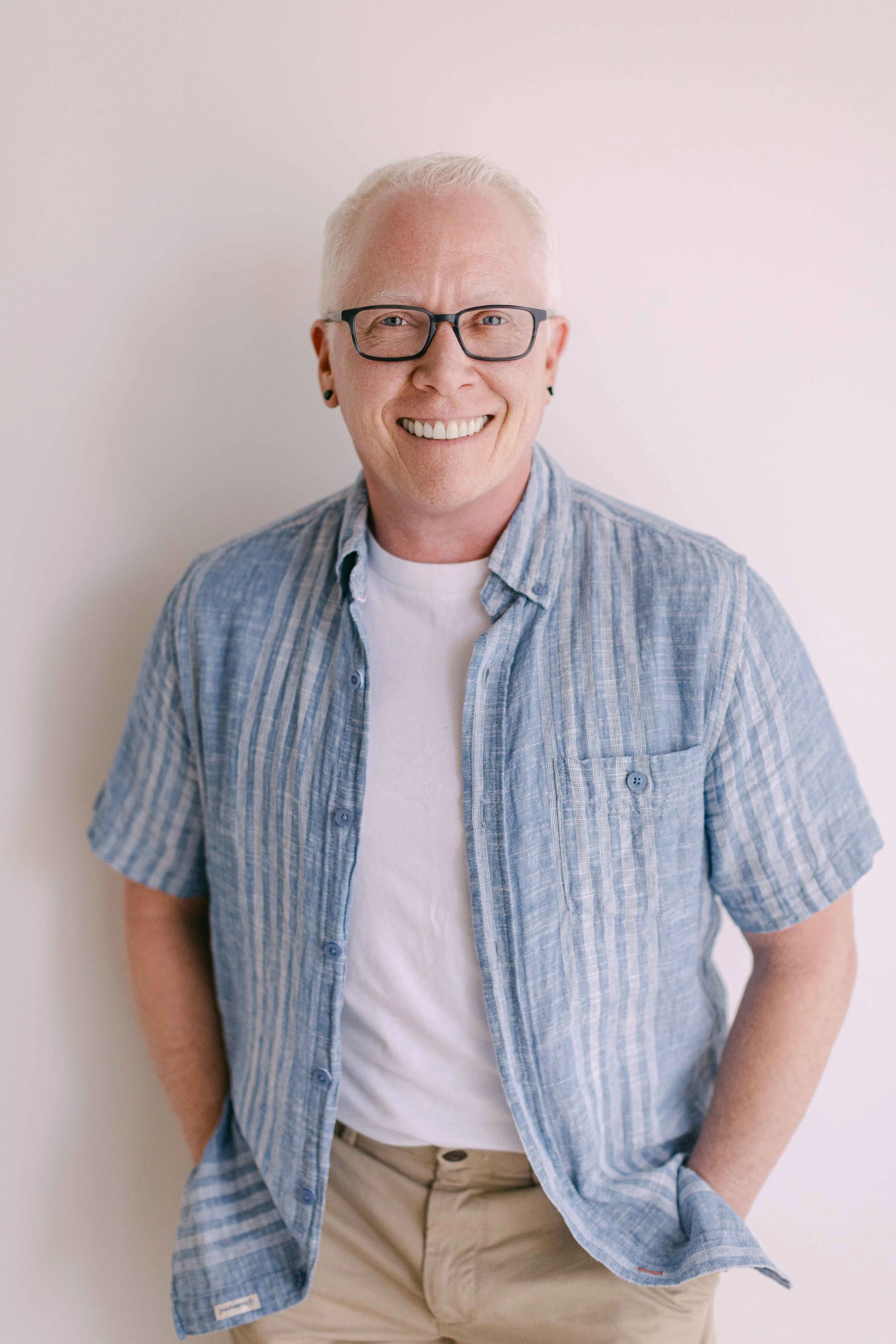A smiling man with blonde hair, glasses, and earrings, wearing a blue checkered shirt over a white T-shirt, and beige pants, standing against a white background.