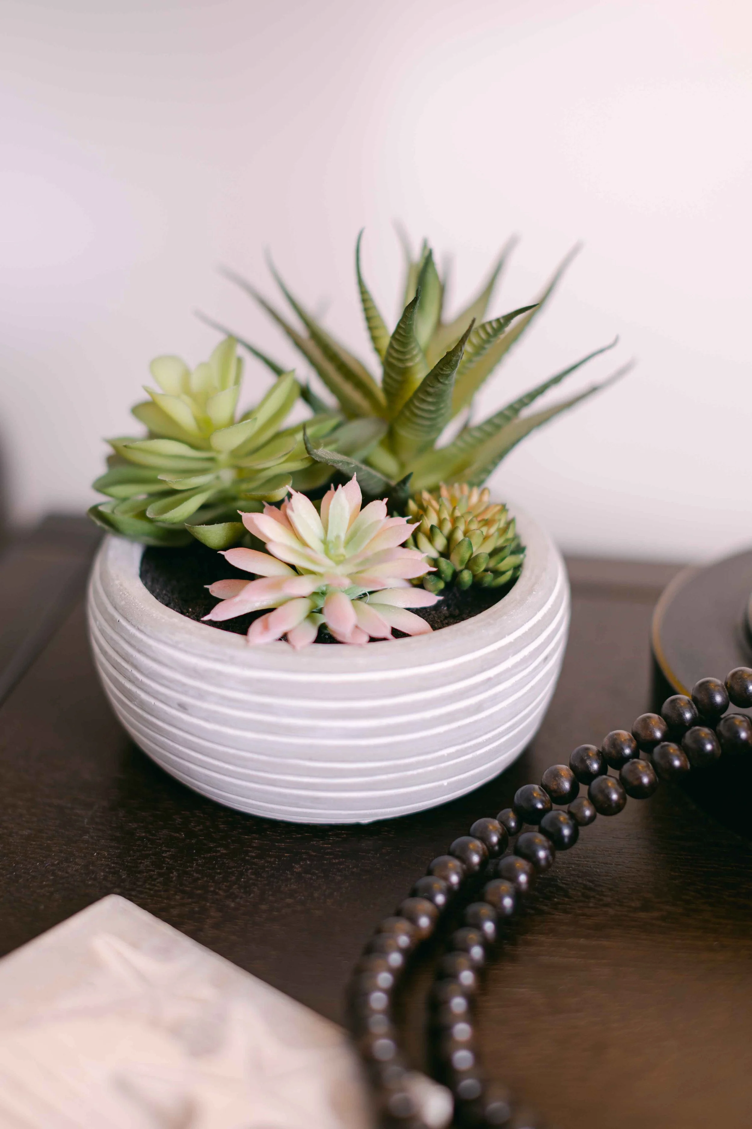 A white round pot with horizontal ridges holding various green and pink succulents, placed on a dark surface with a black beaded necklace nearby.