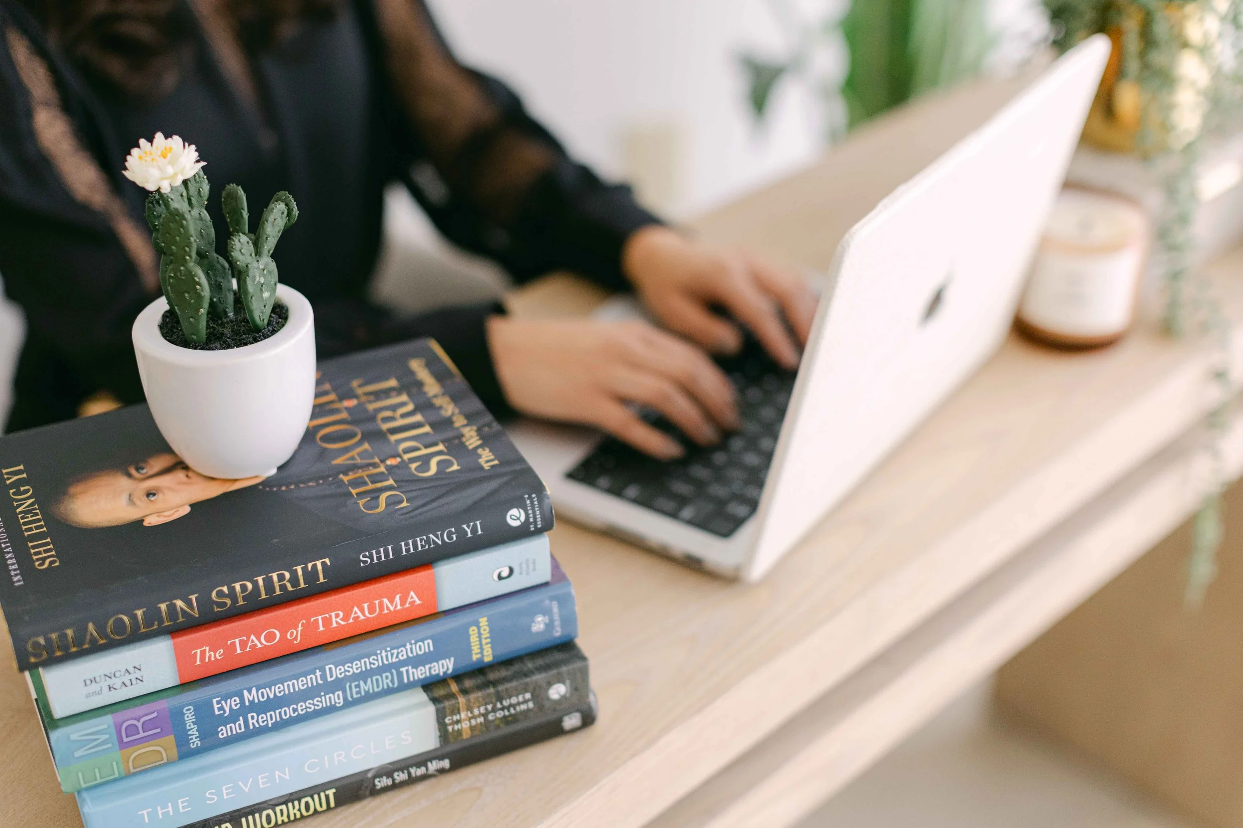 Stacked books on psychology, trauma, eye movement techniques, and mindfulness, with a potted cactus plant nearby, on a wooden surface next to a person typing on a laptop.