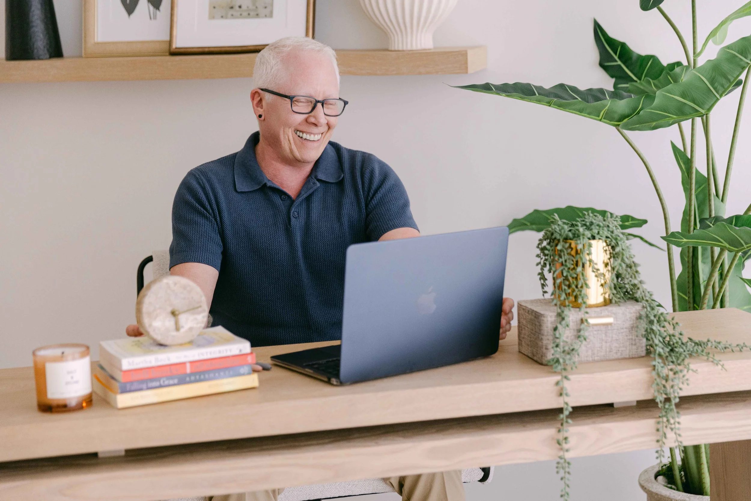 A smiling person with short white hair, glasses, and a dark blue polo shirt sitting at a desk with a laptop, a stack of books, a candle, and a decorative object on it. The background includes framed pictures, a large white vase, and green indoor plants.
