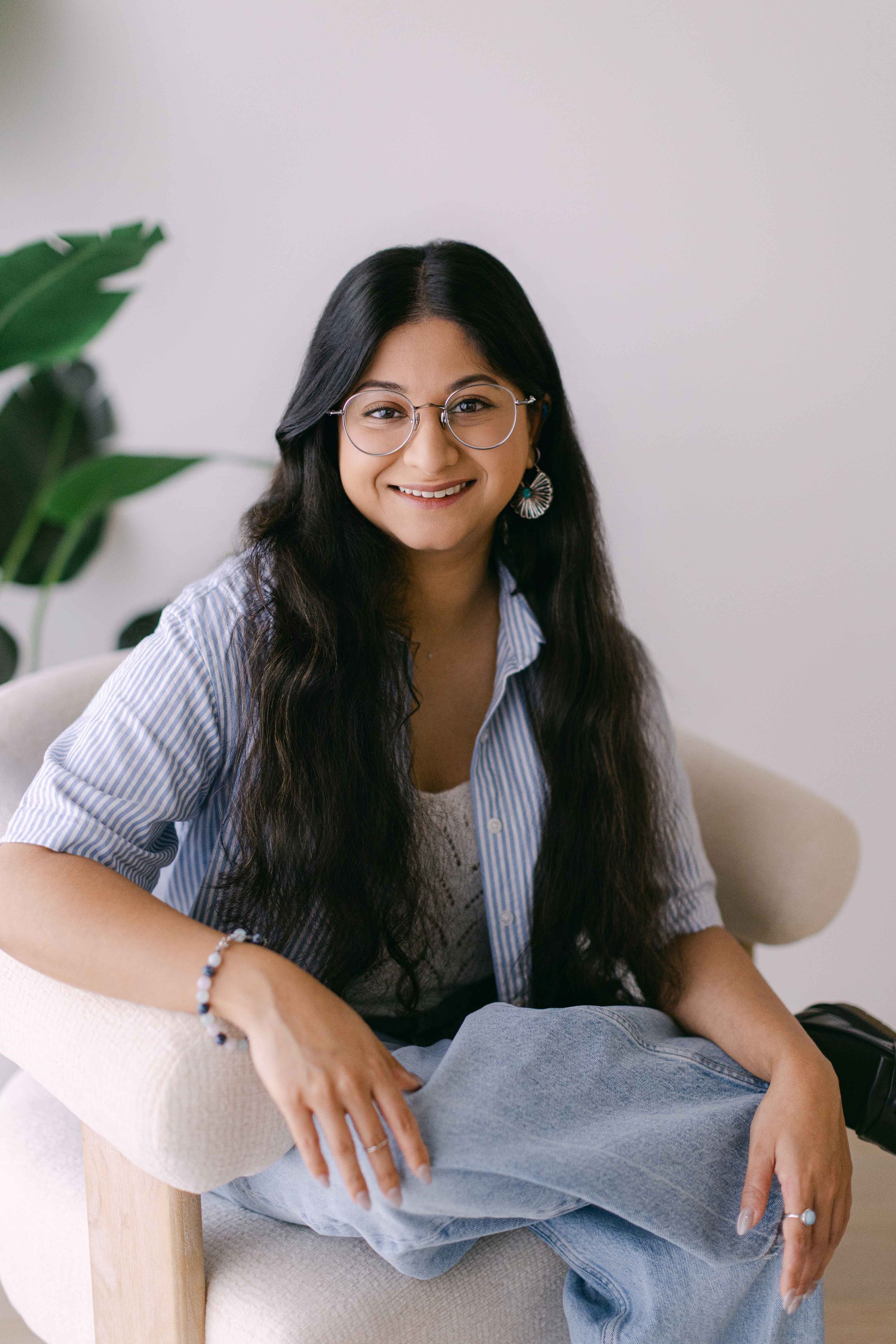 A young woman with long dark hair, glasses, and earrings, smiling and sitting in a cozy chair with her legs crossed. She is wearing a striped shirt and light blue jeans. There's a green plant in the background and a plain white wall.