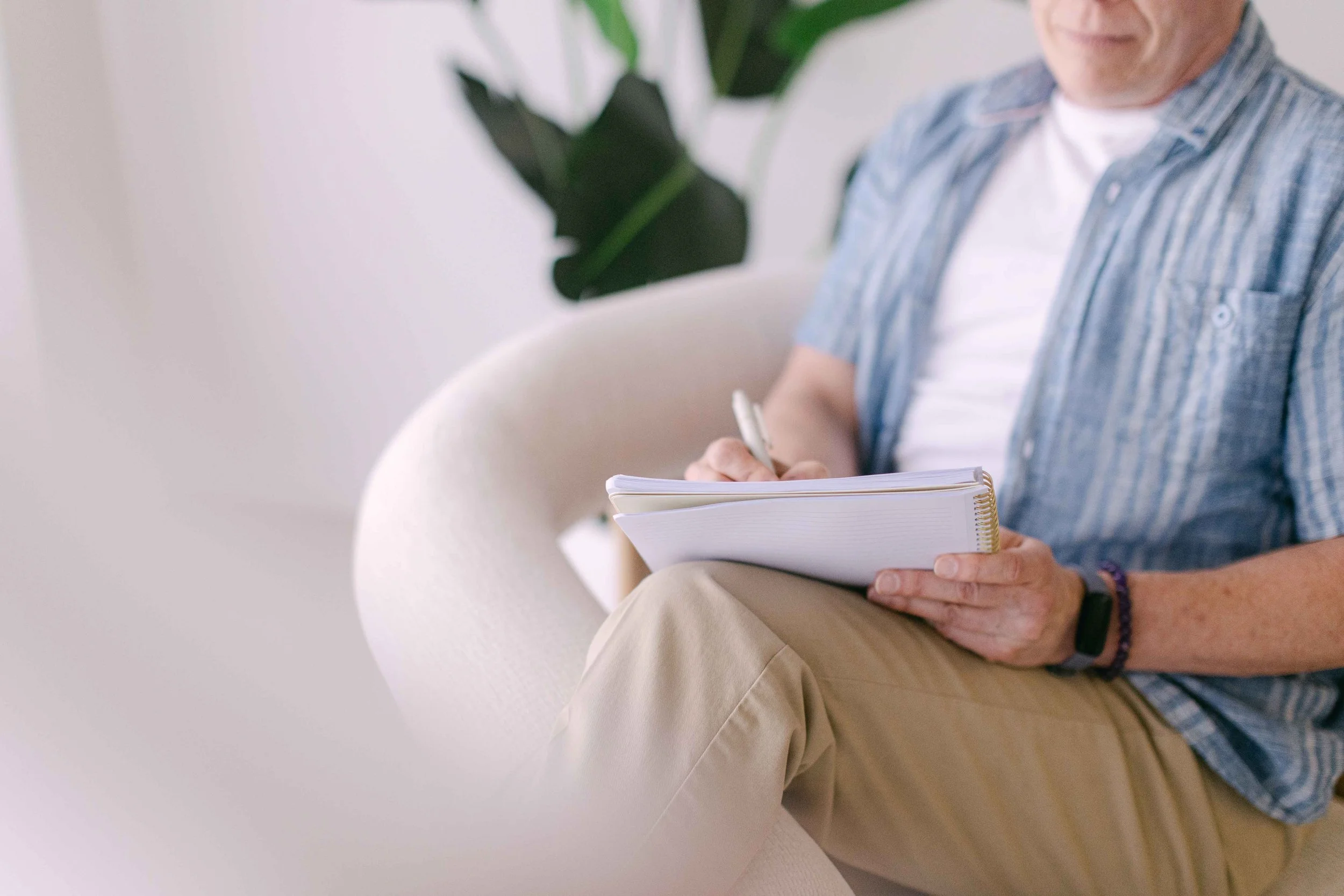 Close-up of a person sitting on a white chair, writing in a notebook with a pen, wearing a plaid shirt and beige pants, with a green plant in the background.
