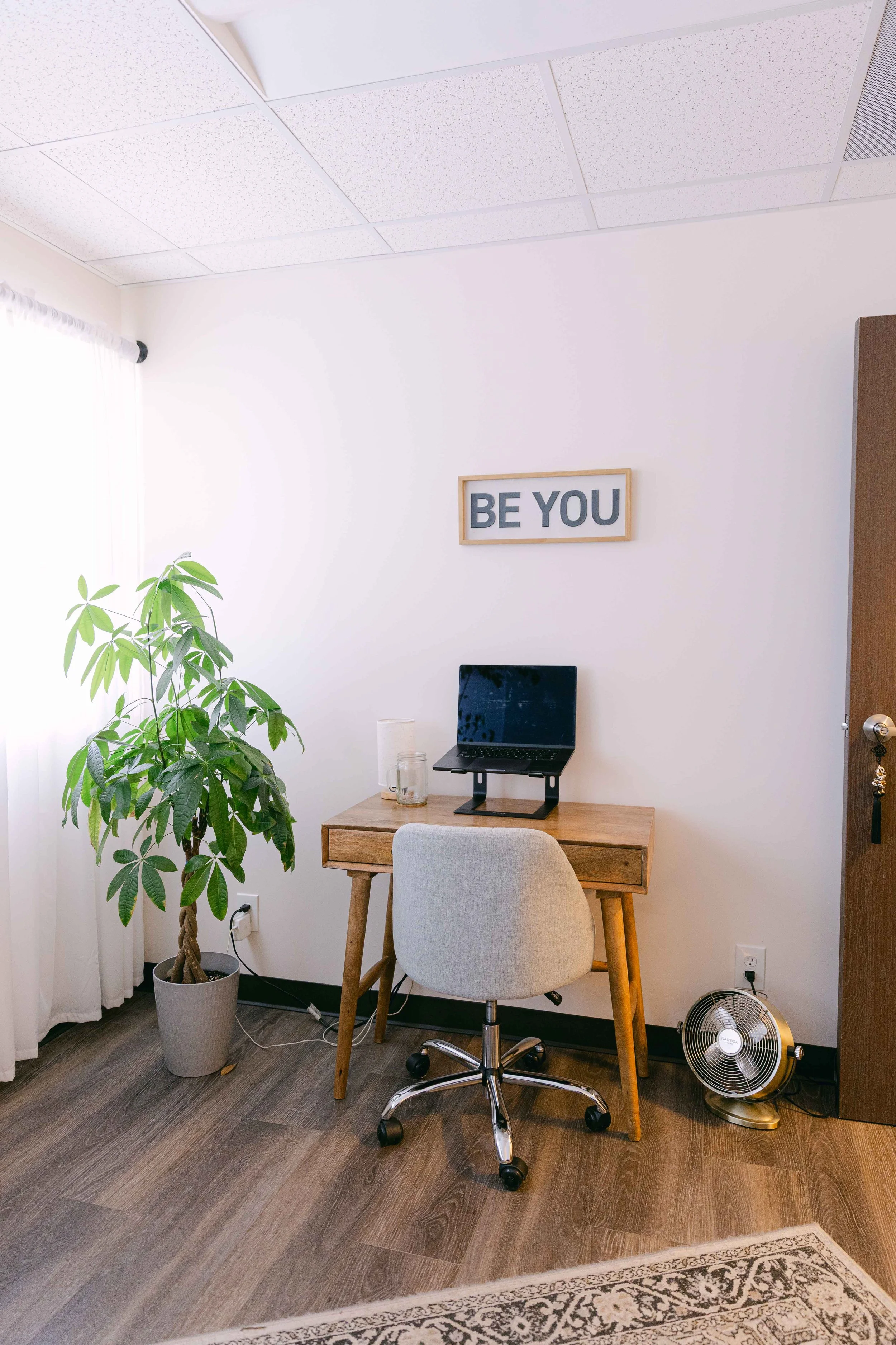 an office workspace with a wooden desk, beige chair, laptop, potted plant, wall sign that says 'BE YOU', and a small fan on the floor