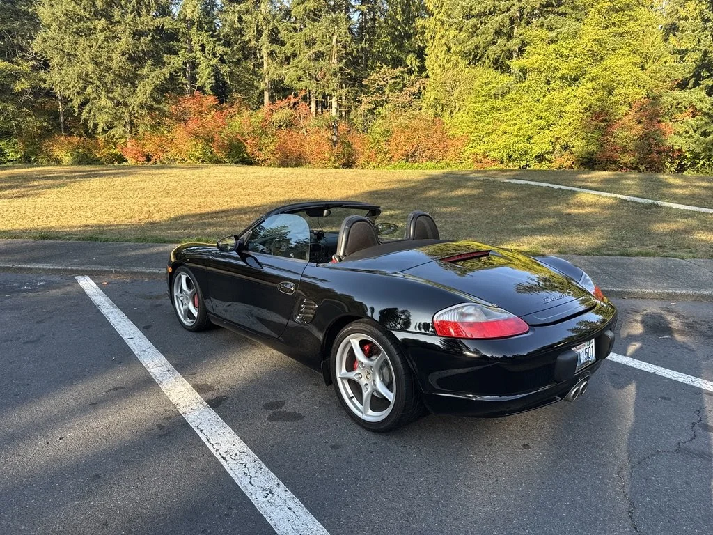 A black Porsche Boxster sports car parked in an empty parking lot with trees and fall foliage in the background.