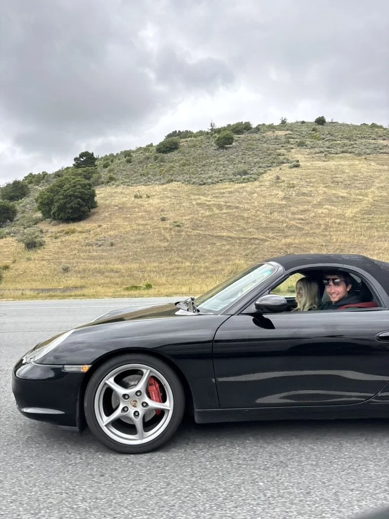 A black convertible sports car with two people inside, driving on a highway with a grassy hillside and cloudy sky in the background.