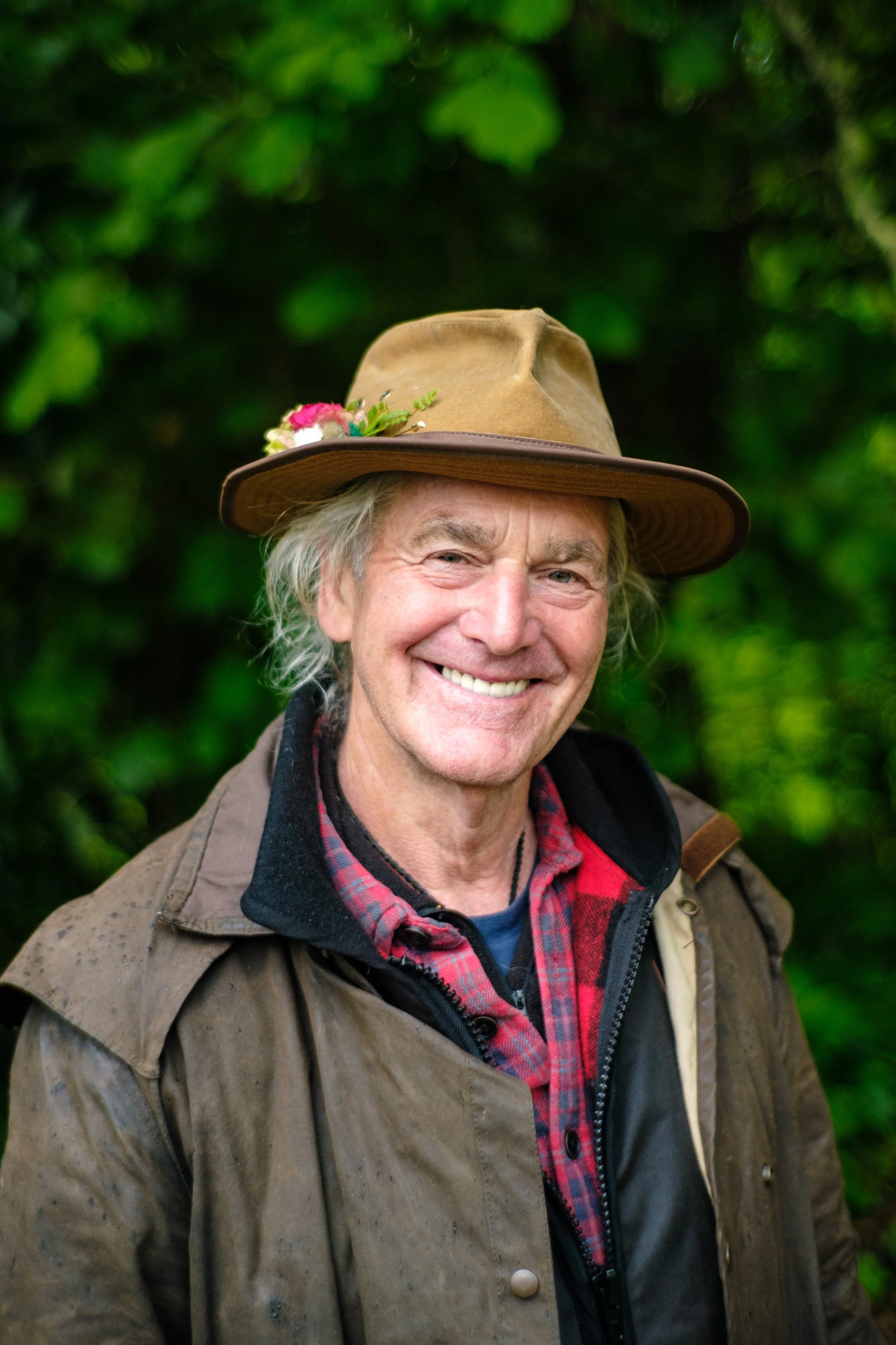 Photo of Charley Bradley smiling into the camera, with a rimmed brown hat and oilskin jacket. Greenery in the background.