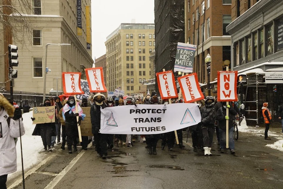 Protest march on a city street with people holding signs and a large banner that reads "Protect Trans Futures". Signs include messages such as "Trans Kids Matter," "Care," "Shelter," and "Now." Snow is on the ground, and the participants are dressed warmly.