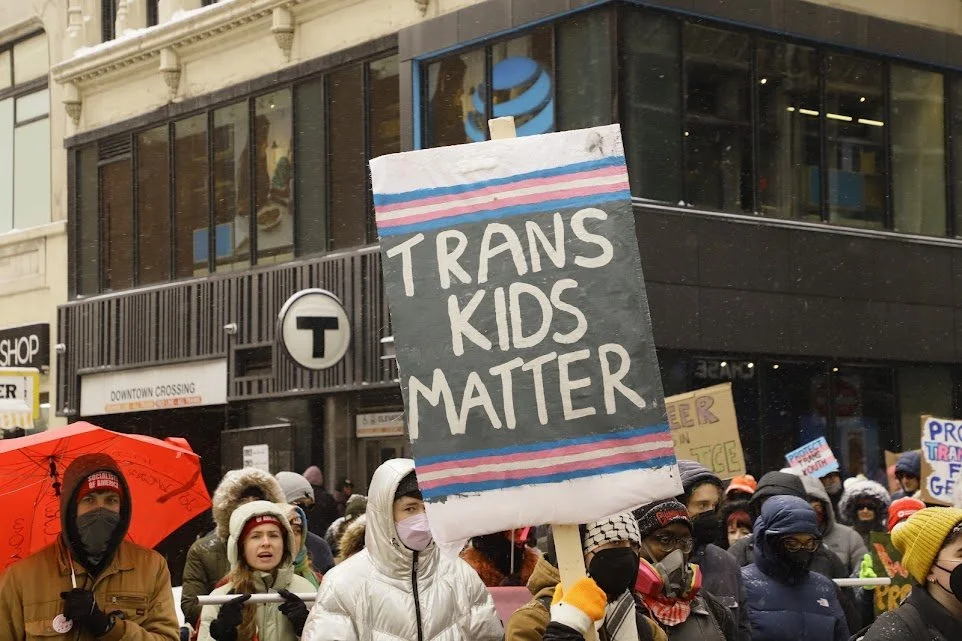 A protest march in an urban area with many people holding signs advocating for trans rights. The central sign reads "Trans Kids Matter" in large white letters on a background with pink, blue, and gray colors. The crowd is diverse, wearing winter clothing and face masks, and the background features a building with a large glass window and a 'T' sign indicating a transit station.