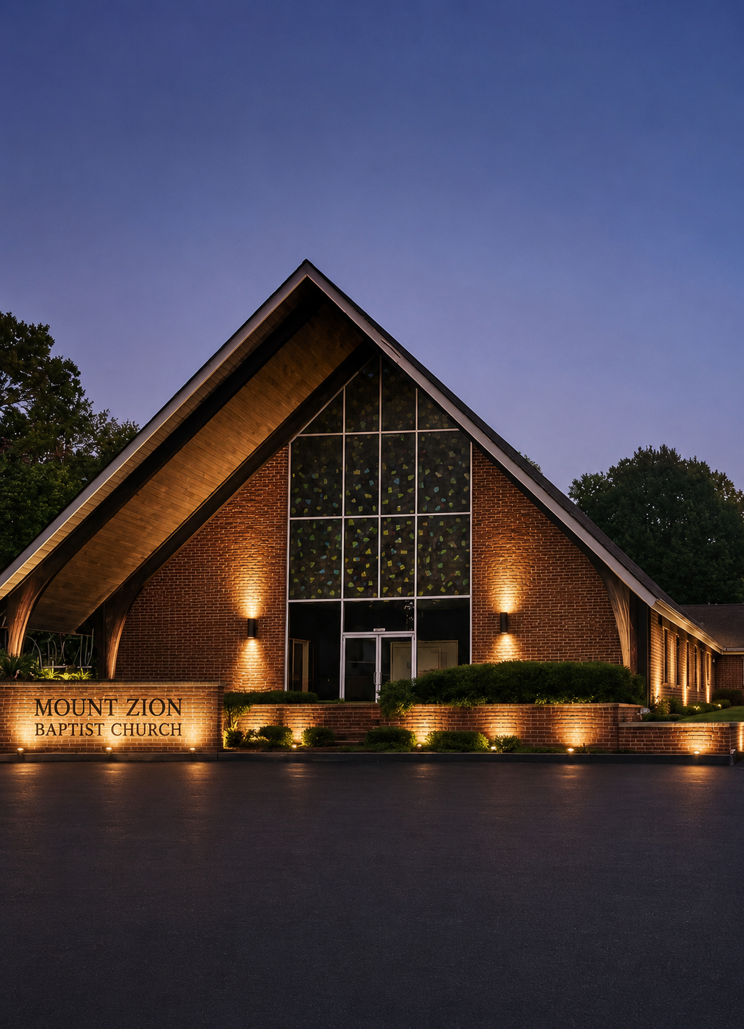 The illuminated exterior of Mount Zion Baptist Church at night, with a large, A-frame wooden and brick structure, stained glass windows, and landscaping in front.