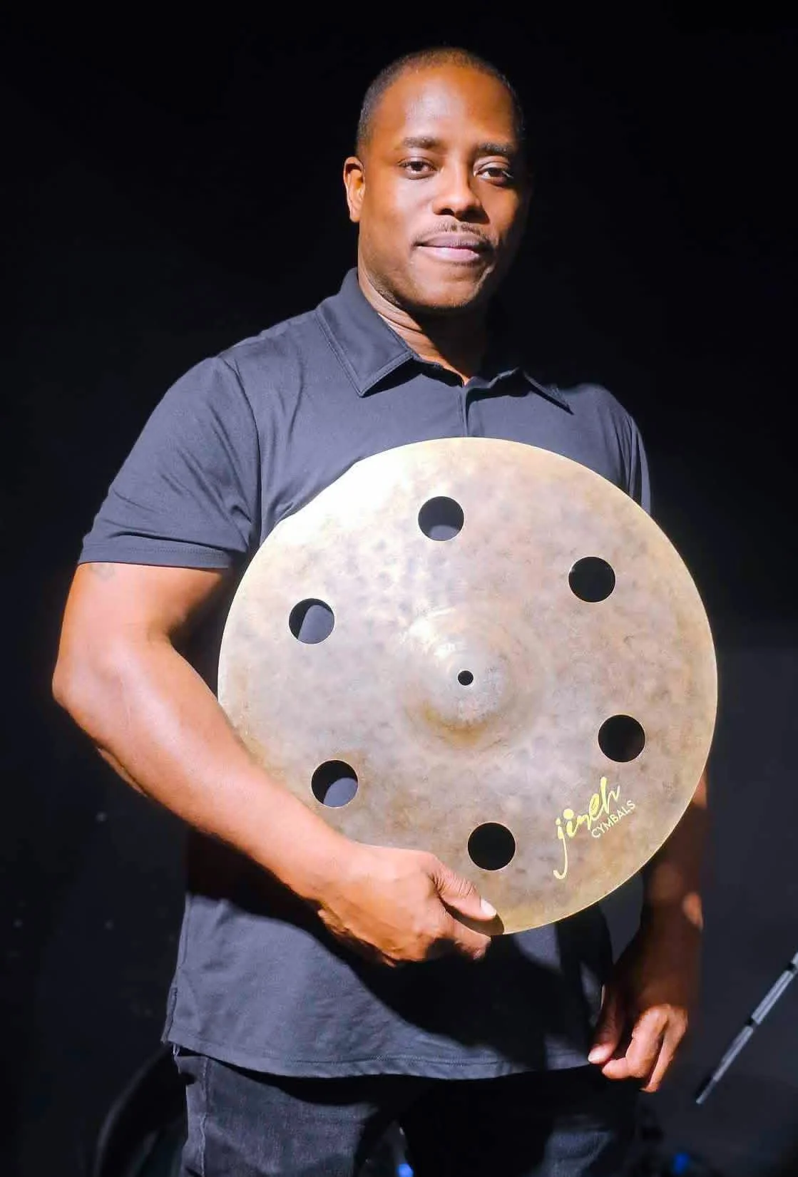 A man holding a large cymbal with the brand name 'Jemel' printed on it, standing against a black background.