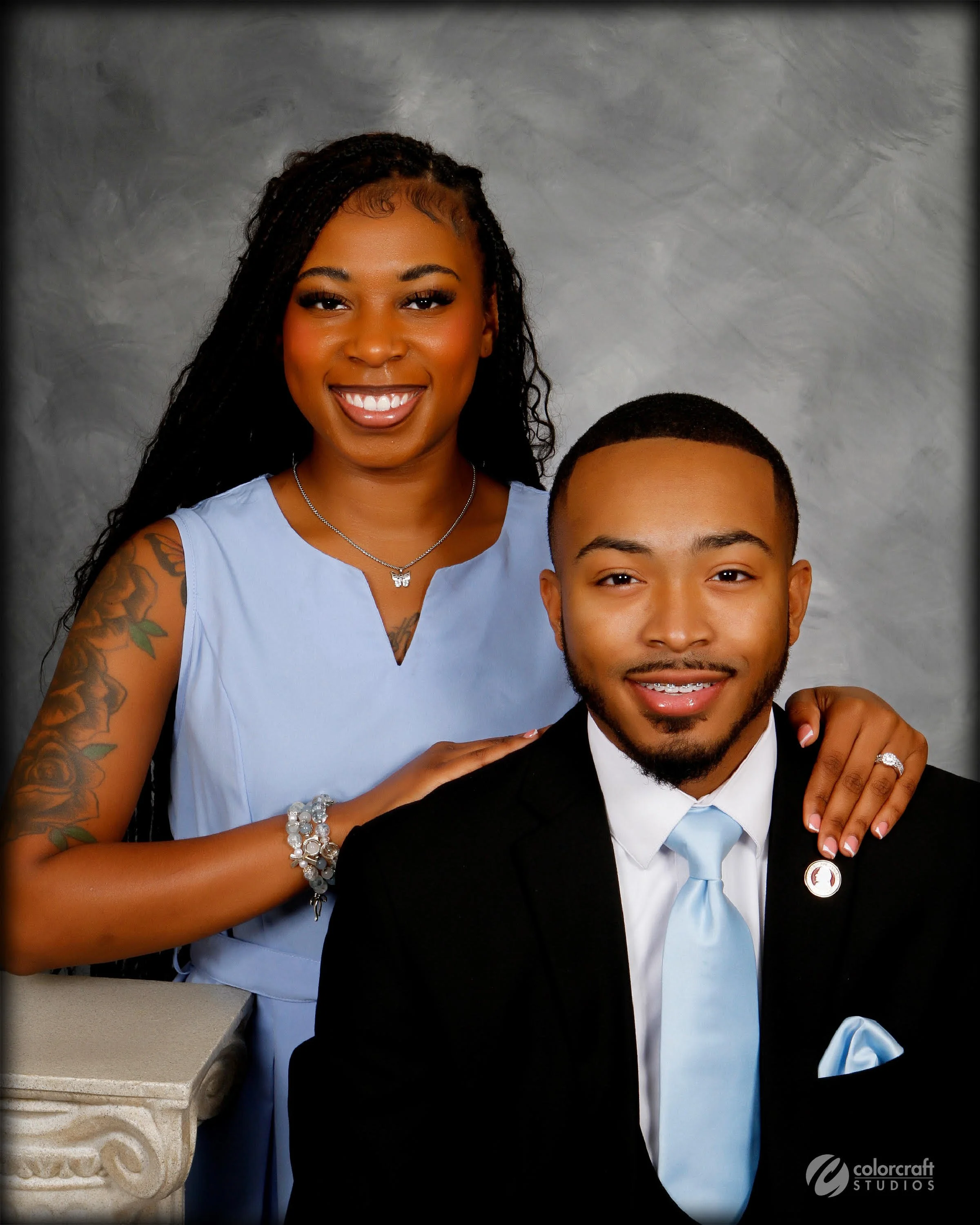 A smiling woman with long braided hair, wearing a light blue dress and a butterfly necklace, standing beside a young man in a black suit and light blue tie, against a gray textured background.