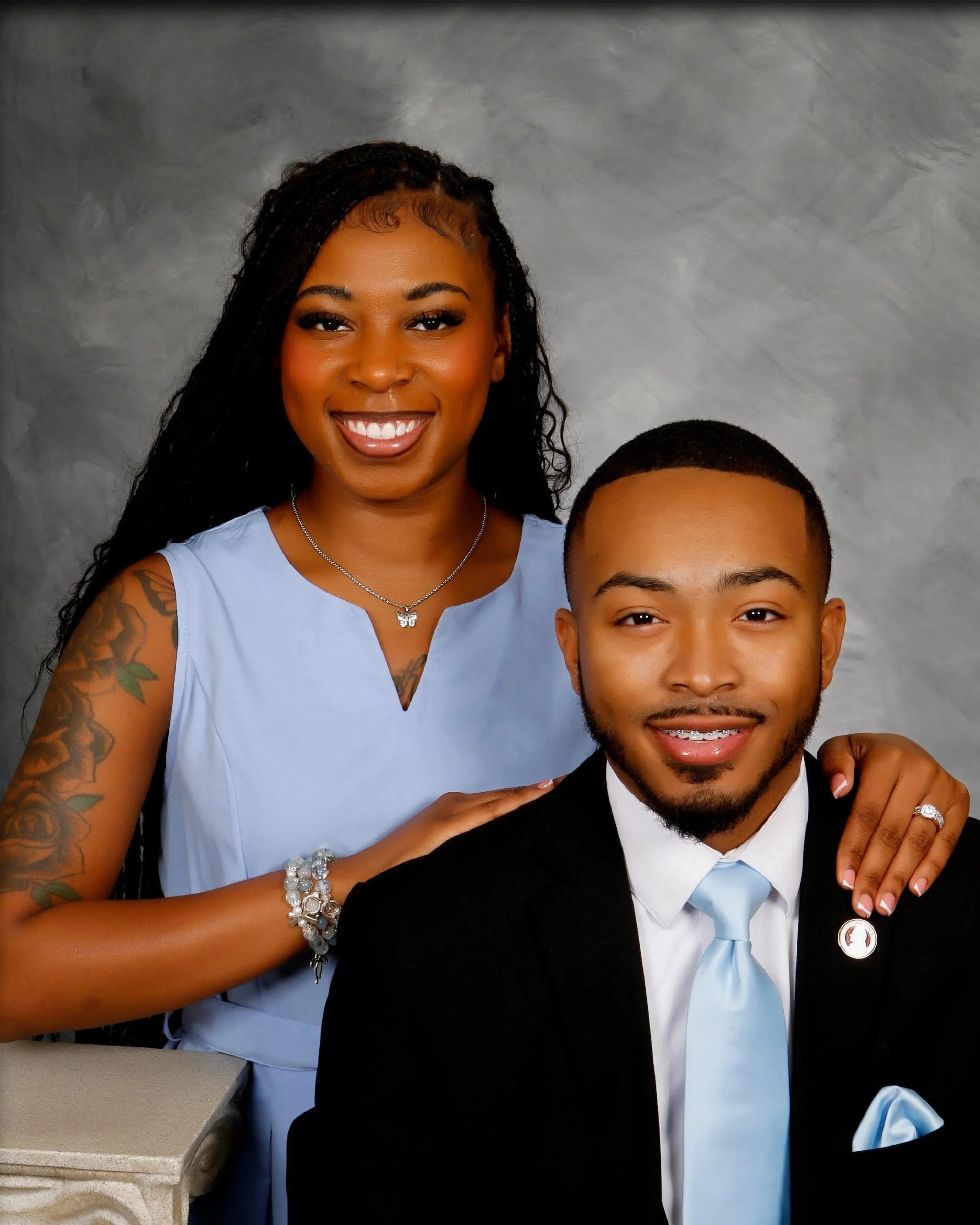 A woman with long curly hair, wearing a light blue dress, silver jewelry, and a butterfly necklace, stands next to a young man in a black tuxedo with a light blue tie, both smiling at the camera against a gray background.