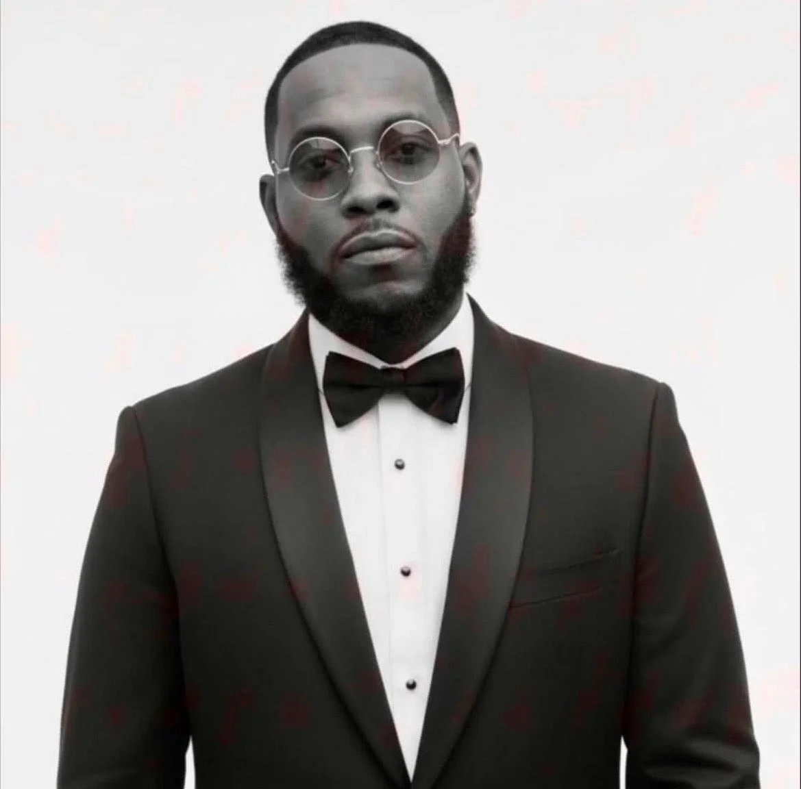 A man wearing a black tuxedo with a bow tie, round glasses, and a groomed beard, standing against a plain white background.
