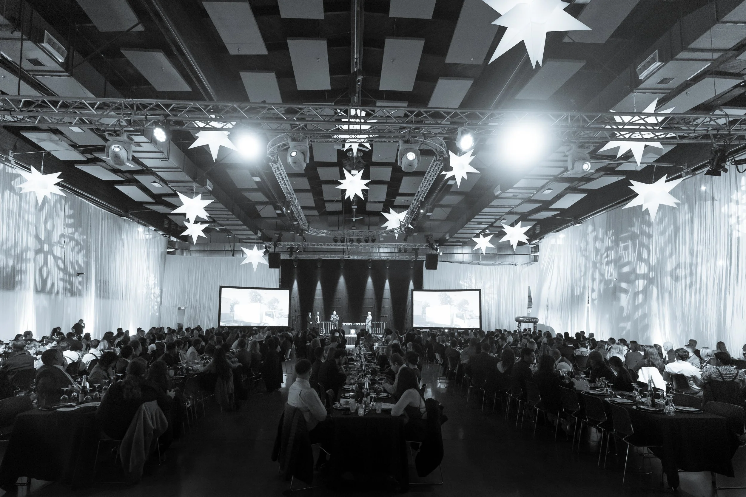 Large conference or event hall filled with seated attendees listening to a presentation on stage with large screens. The ceiling has decorative star-shaped lights and stage lighting.
