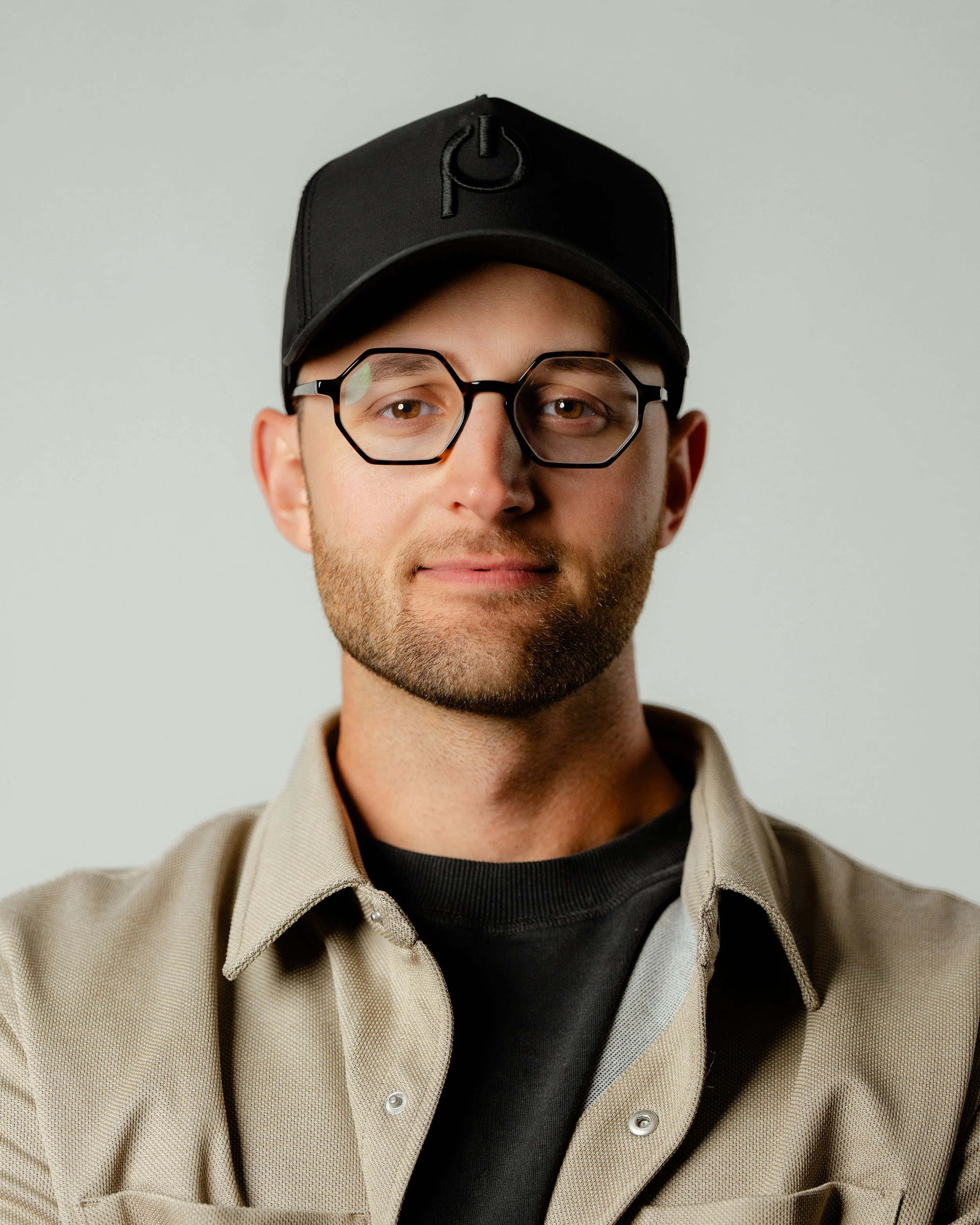 A young man with a beard and glasses wearing a black cap, black shirt, and beige jacket against a plain light gray background.